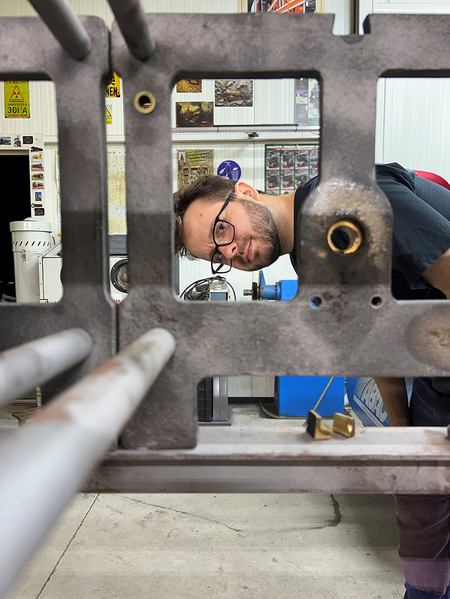 Na fotografii widzimy fragment dużego mechanizmu zegara wieżowego w pracowni konserwatorskiej. Pierwszy plan niemal w całości zajmuje masywna, ciemnoszara metalowa rama mechanizmu, z prostokątnymi otworami, tulejami i wystającymi wałami.
Przez jeden z dużych otworów w ramie zagląda mężczyzna. Ma krótkie ciemne włosy, zarost i czarne okulary. Pochyla głowę na bok i spogląda w stronę aparatu, dzięki czemu jego twarz zostaje niejako „oprawiona” przez prostokątny prześwit konstrukcji zegara.
Metalowa rama nosi ślady użytkowania i prac konserwatorskich. Jej powierzchnia jest matowa, miejscami nierówna i przebarwiona. W kilku miejscach widoczne są mosiężne tuleje i drobne elementy montażowe. Po lewej stronie kadru z ramy wystają dwa długie, jasne wałki, mocno rozmyte ze względu na niewielką głębię ostrości.
W tle znajduje się wyposażenie pracowni: urządzenia techniczne, fragmenty narzędzi, plakaty i tablice na ścianach. Kolorystycznie dominują szarości metalu, biel ścian oraz pojedyncze niebieskie elementy wyposażenia.
Fotografia ma nieco żartobliwy, bezpośredni charakter. Pokazuje skalę mechanizmu zegara wieżowego w zestawieniu z człowiekiem i jednocześnie pozwala zajrzeć do jego masywnej, otwartej konstrukcji.