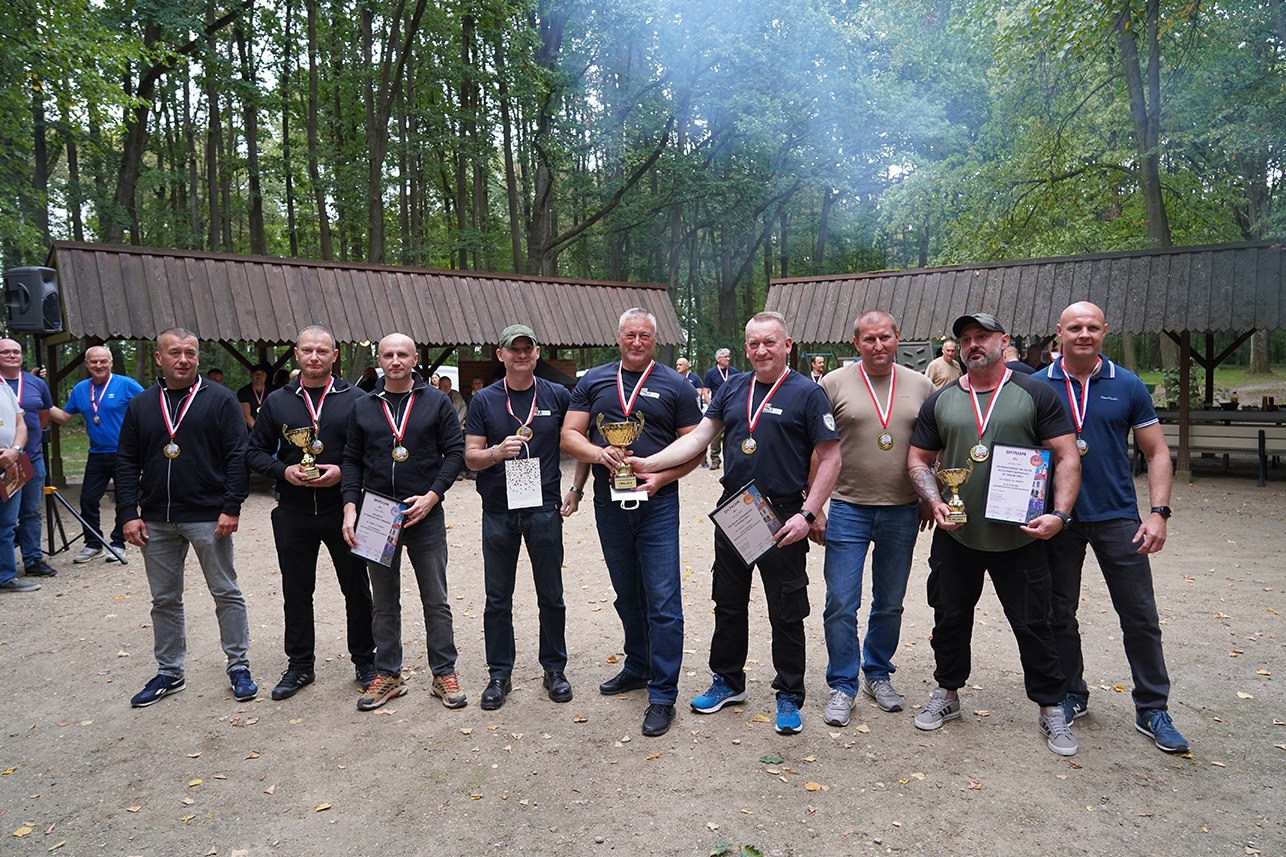 Na terenie miejsca ogniskowego w Muzeum Zamoyskich w Kozłówce stoi dziewięciu zdobywców trzech pierwszych miejsc w IX Turnieju Wewnętrznych Służb Ochrony. Pierwsze miejsce zajęła drużyna Muzeum Zamkowego w Malborku, drugie – Muzeum w Nieborowie i Arkadii, a trzecie – Państwowego Muzeum Auschwitz-Birkenau w Oświęcimiu. Mężczyźni pozują z medalami, pucharami, dyplomami i nagrodą. W tle pozostali uczestnicy wydarzenia.