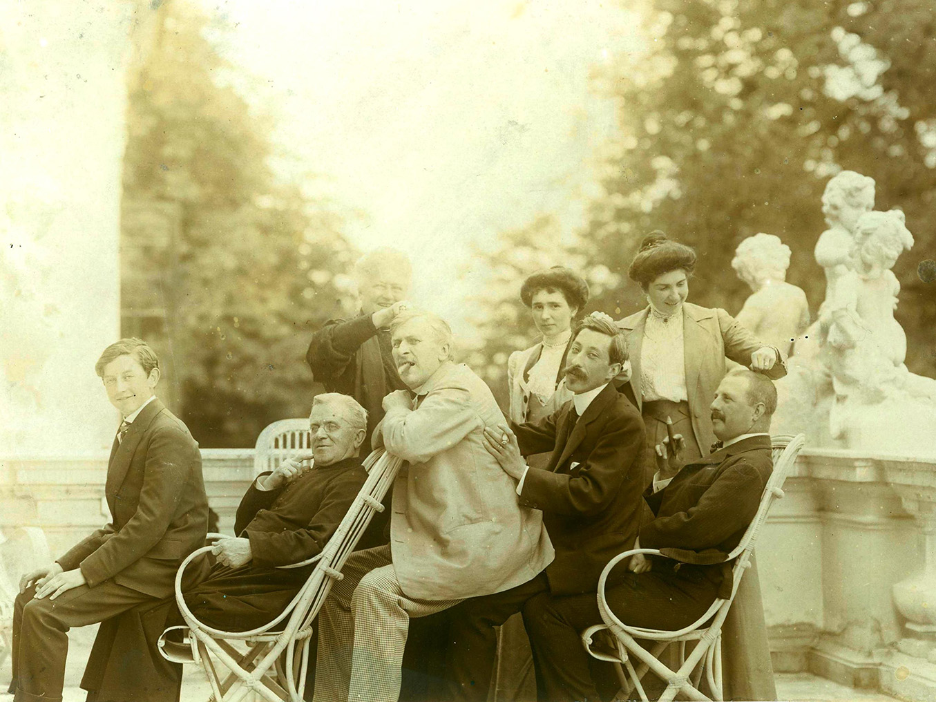 Stara, pożółkła fotografia z początków XX wieku, utrzymana w żartobliwym tonie. Grupa osób na tarasie. Pięciu mężczyzn w różnym wieku, siedząc na kolanach poprzednika tworzy „wagoniki”. Za nimi trzy kobiety, dwie młodsze i jedna starsza, chwytają dłonią za ich włosy na czubku głowy.