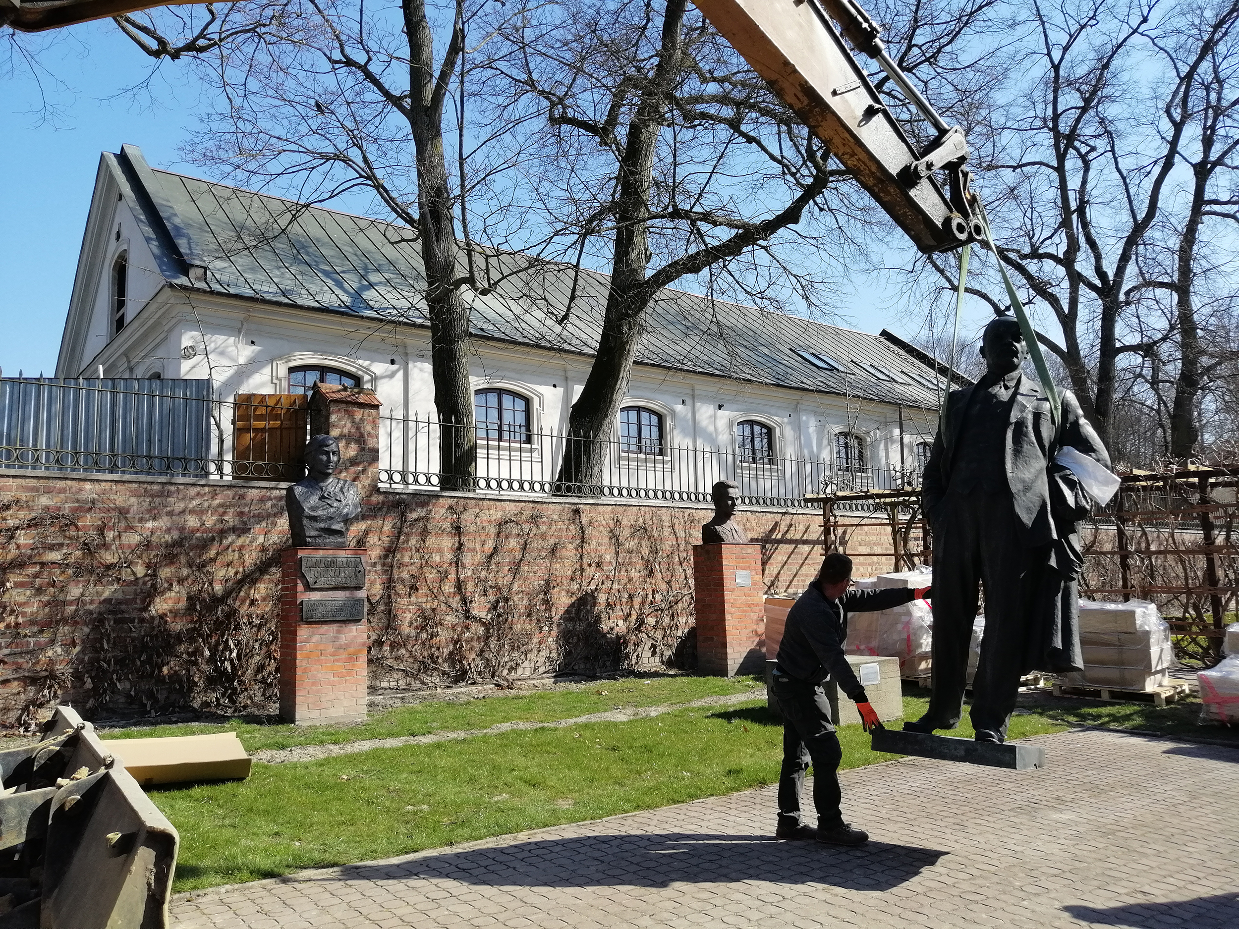 Fotografia. Pomnik Włodzimierza Lenina, postać całościowa. Rzeźba z brązu o wymiarach 260 x 120 x 75 cm uniesiona nad ziemię za pomocą pasów holowniczych. Po lewej stronie mężczyzna podtrzymujący rzeźbę.