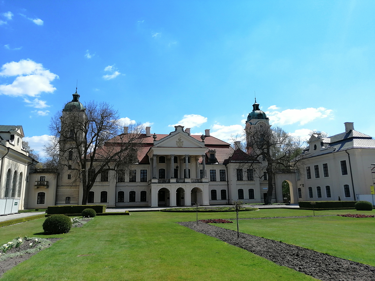 Na tle błękitnego nieba okazały piętrowy pałac pokryty dwuspadowym dachem. Po obu stronach rezydencji zakończone kopułą wieże, połączone murkami z przyległymi budynkami. Całość tworzy literę U. Przed pałacem reprezentacyjny dziedziniec we wczesno wiosennej odsłonie.