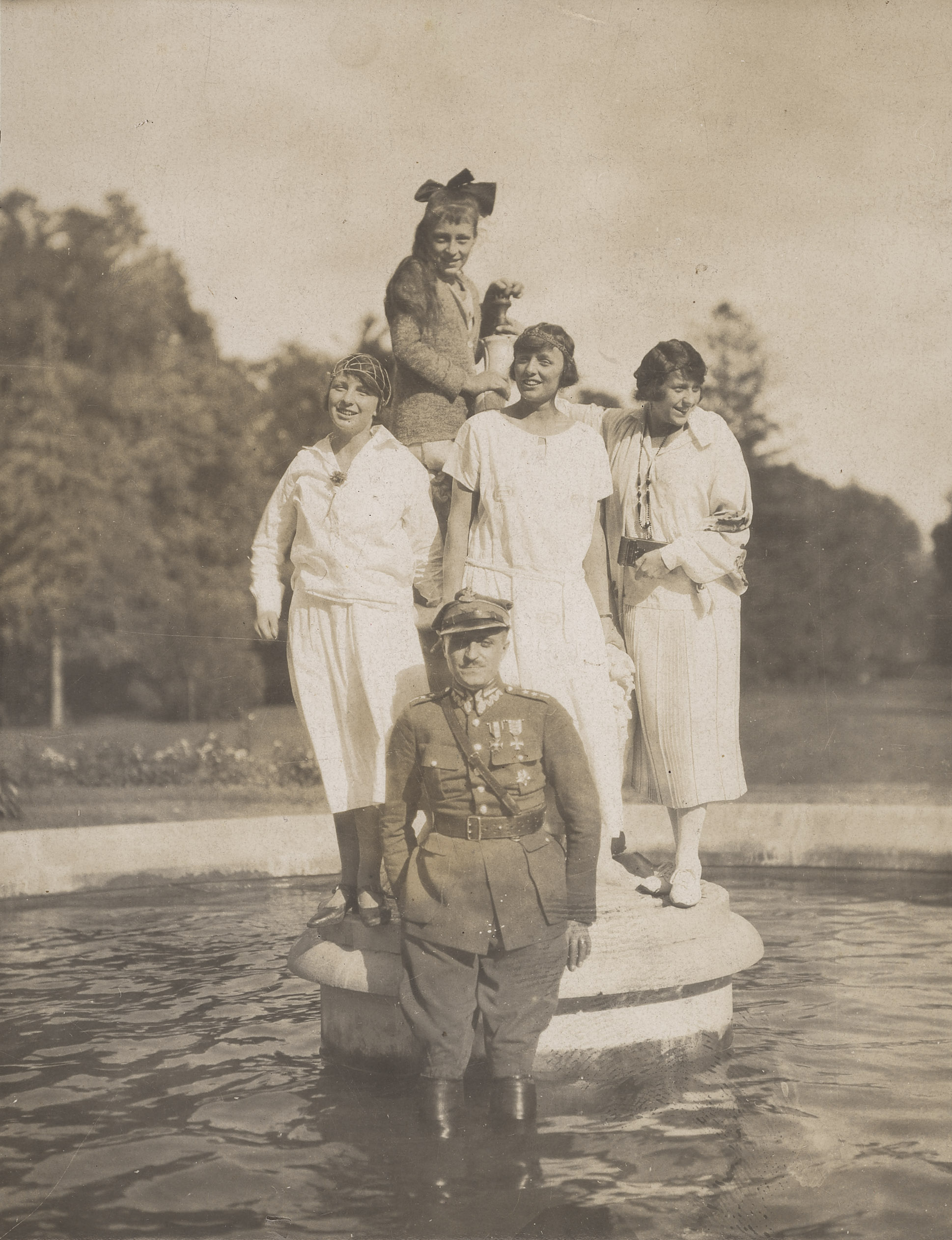 Aleksander Zamoyski z żoną Jadwigą z Brzozowskich (pierwsza z prawej) oraz jej siostrami (od lewej: Izabelą, Różą i Marią. Czarno-biała fotografia. Aleksander Zamoyski z żoną Jadwigą i jej siostrami: Izabelą, Różą i Marią. Młodzi ludzie w fontannie. Na pierwszym planie mężczyzna ubrany w mundur stoi w wodzie po kolana. Za nim, stojące na cokole fontanny, trzy kobiety ubrane w jasne, proste sukienki z niskim stanem. Włosy krótkie, ozdobione delikatnymi dodatkami. Za nimi, stojąca najwyżej najmłodsza kobieta ubrana w sweter. W długie włosy wpięta na czubku głowy duża kokarda.