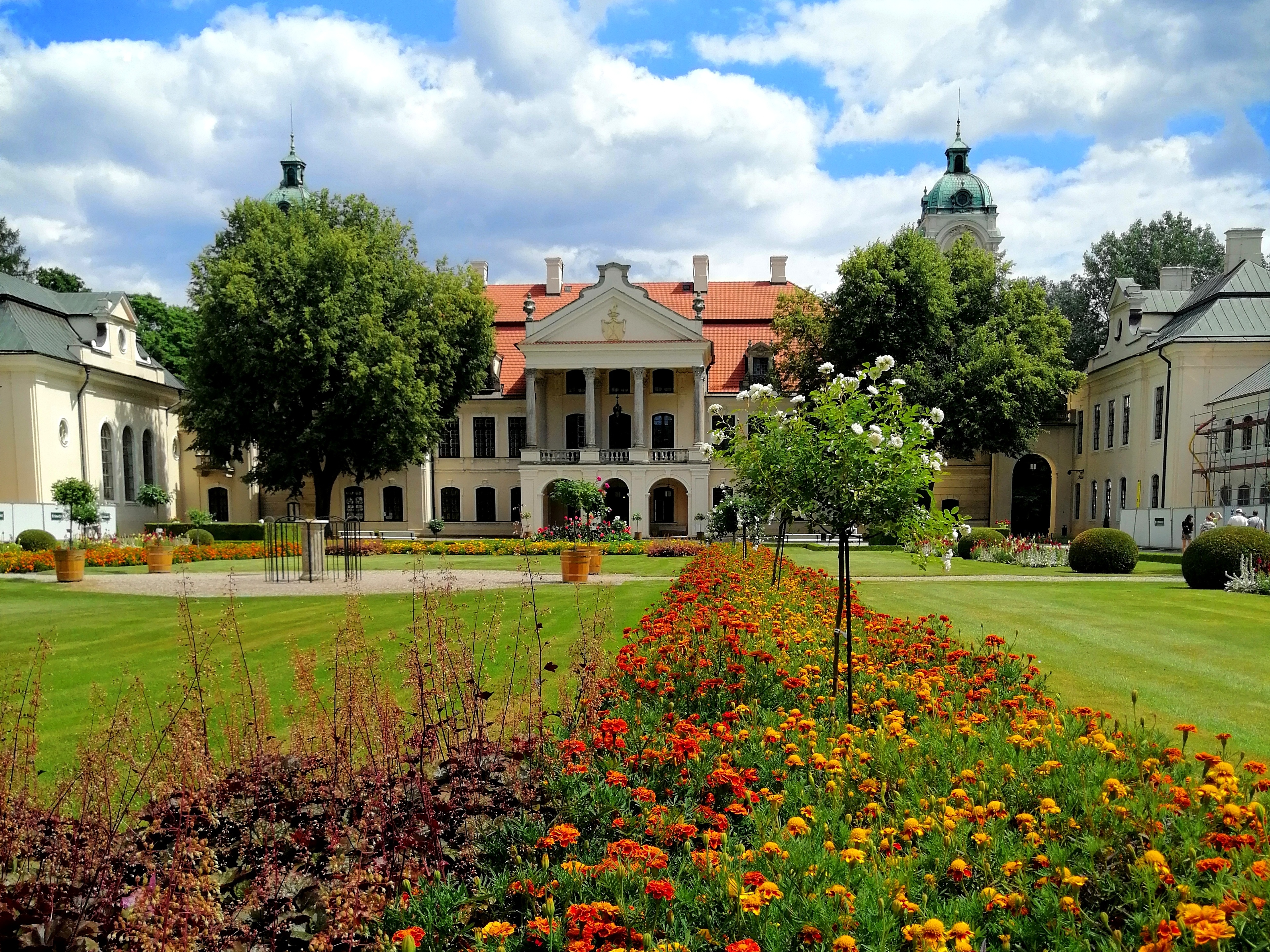 Fotografia. Na pierwszym planie kwitnące kolorowe, drobne kwiaty. W tle okazały piętrowy budynek z wysuniętym portykiem, pokryty mansardowym dachem z czerwoną dachówką. Po obu stronach pałacu, ustawione prostopadle dwa wysokie budynki z jasną elewacją i zielonym dachem. Przed pałacem dwa wielkie liściaste drzewa.