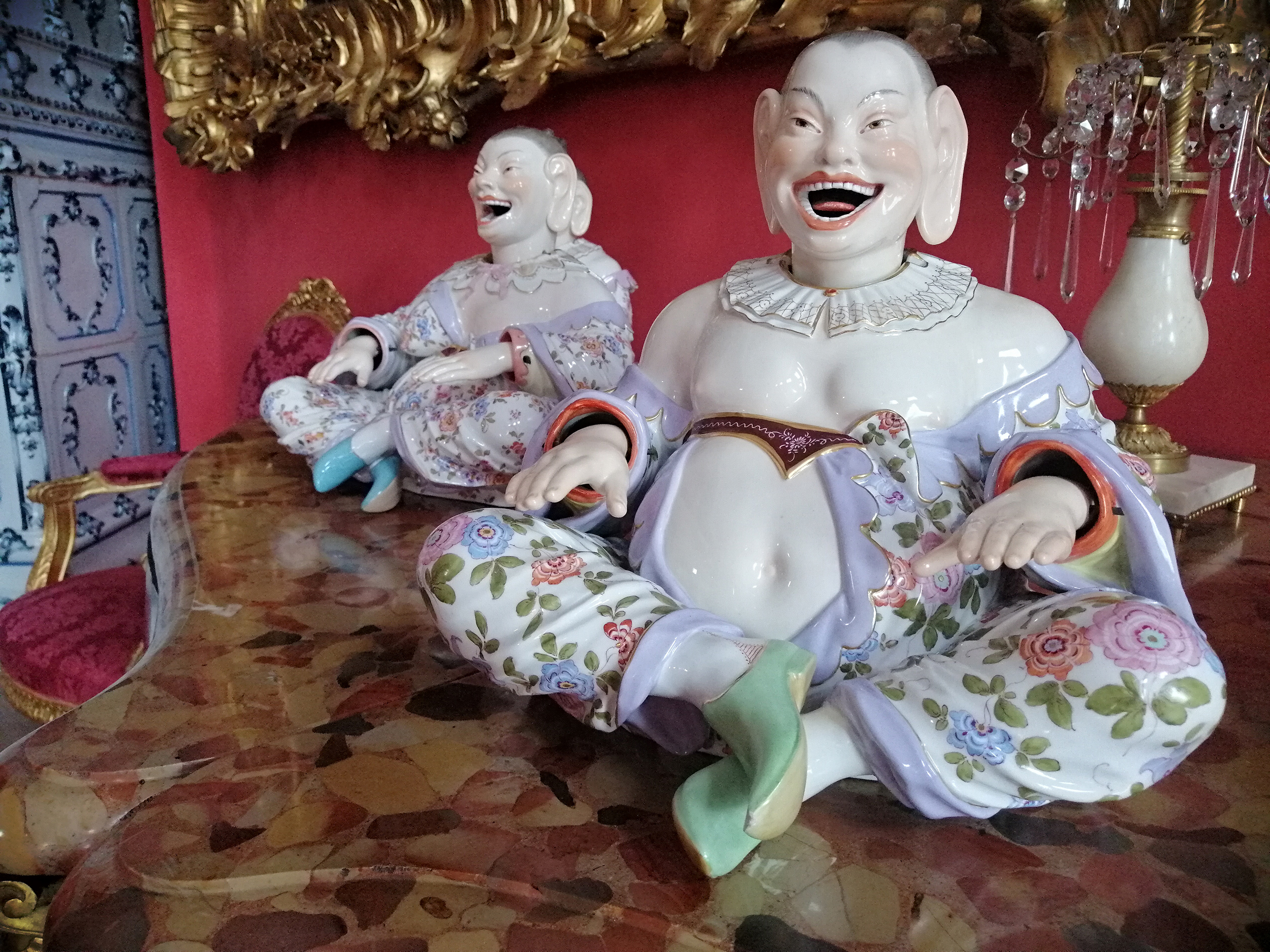 Fotografia. Na pierwszym planie porcelanowa figurka pulchnego Chińczyka siedzącego na marmurowym blacie. Głowa i dłonie stanowią oddzielne, ruchome elementy. Twarz pyzata z zarysowanymi rumieńcami. Oczy skośne z wyrysowanymi w cienkie łuki brwiami. Usta czerwone, w szczerym uśmiechu odsłaniającym białe zęby i język. Duże uszy. Przy szyi kołnierzyk. Ubrany niedbale w kwieciste kimono z liliową lamówką, odsłaniające ramiona i duży brzuch. Na stopach zielone wsuwane pantofle. Po lewej stronie, lekko w głębi, analogiczna figurka.