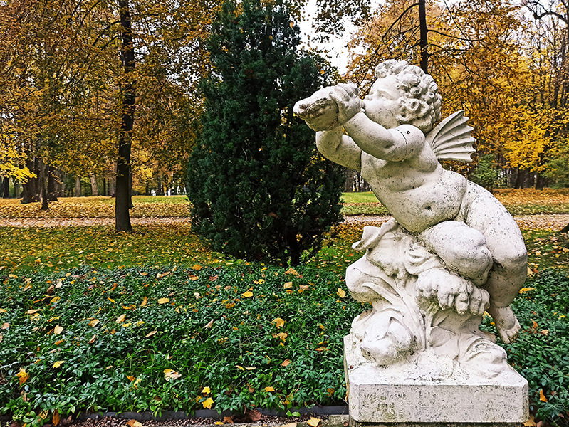 Fotografia pozioma. Barokowa rzeźba plenerowa przedstawiająca putto – nagiego, pulchnego chłopca. Dziecięca postać siedząca na cokole stworzonym z fal i morskich roślin, skręcona ekspresyjnie w lewą stronę. Na głowie burza loków. Twarz życzliwa, pyzata. Nadęte policzki dotykają muszli trzymanej w wyciągniętych do przodu rączkach.  Przy pleckach mała płetwa. Na cokole, po lewej napis Val d’Osne Paris.