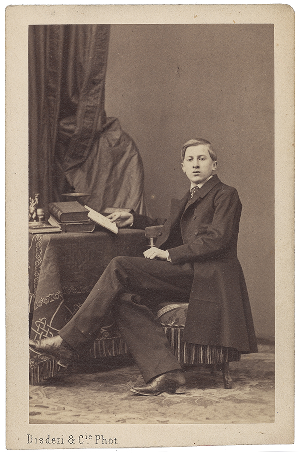 Fotografia zabytkowa, pionowa. Młodzieniec w całej postaci, siedzący przy stoliku, zwrócony w lewo, głowa en face; w tle z lewej kotara.