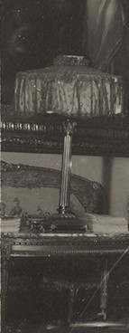 Lampa naftowa, kolumnowa, Salon Biały, Kozłówka, 1929 rok; fragment fotografii archiwalnej; zbiory Muzeum Zamoyskich w Kozłówce, nr inw. MPK/F/1932 Fotografia archiwalna czarno biała fotografia przedstawiająca lampę ustawioną na stole we wnętrzu pomieszczenia. Lampa ma kolumnową podstawę i tekstylny szeroki i płaski abażur.