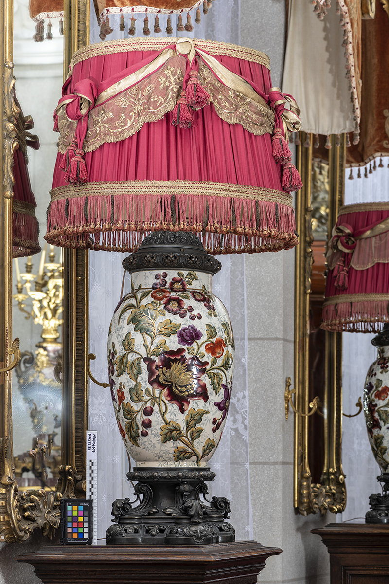 Lampa stołowa, salonowa, Klatka Schodowa pałacu kozłowieckiego; zbiory Muzeum Zamoyskich w Kozłówce, nr inw. MPK/CS/308 Fotografia przedstawia lampę o korpusie w kształcie wazy, na niej namalowany duży kwiatowy wzór. Podstawa lampy ciemna, dekorowana. Szczyt lampy okryty abażurem w kształcie ściętego stożka. Czerwona tkanina abażuru udekorowana wstążkami i frędzlami. W tle duże lustra w złoconej ramie.