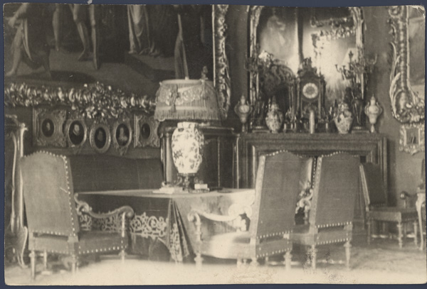 Salon Czerwony, Kozłówka, przełom lat 30. i 40. XX wieku; fotografia archiwalna; zbiory Muzeum Zamoyskich w Kozłówce, nr inw. MPK/F/1852 Fotografia archiwalna, czarno-biała przedstawia wnętrze pomieszczenia z licznymi obrazami oprawionymi w ramy. Na pierwszym planie stół otoczony fotelami, na nim duża lampa z korpusem w kwiaty i abażurem z tkaniny. W tle kominek, nad nim wysokie lustro.