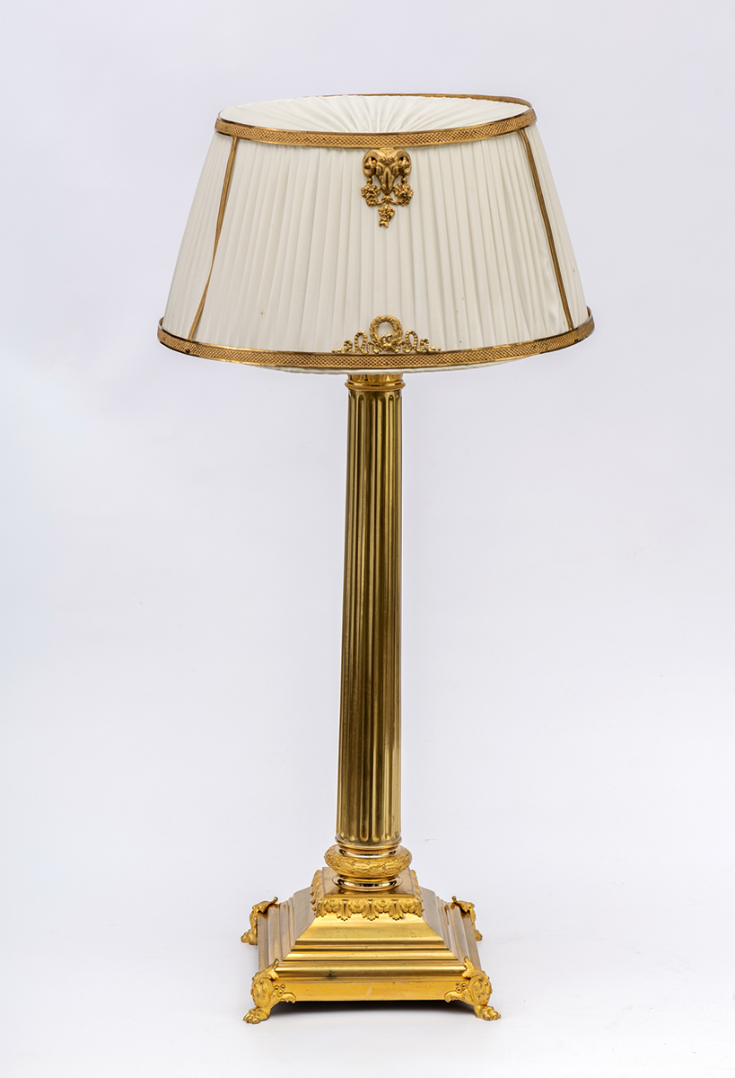 Podstawa lampy stołowej, kolumnowej, A. Robert, Francja, Paryż, przełom XIX/XX wieku, z dobranym ze zbiorów kozłowieckich abażurem, Salon Czerwony pałacu kozłowieckiego; zbiory Zamoyskich w Kozłówce, nr inw. MPK/F/92 Fotografia przedstawia lampę o kolumnowej podstawie w kolorze złotym z jasnym abażurem z tkaniny rozpiętej na dekorowanych obręczach.