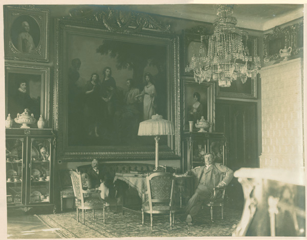 Salon Biały, Kozłówka, przed 1901 rokiem; fotografia archiwalna; zbiory Muzeum Zamoyskich w Kozłówce, nr inw. MPK/F/1875 Fotografia archiwalna w kolorze sepii przedstawia widok wnętrza. W głębi stół z trzema krzesłami i kanapą. Na stole lampa z jasnym abażurem. Na kanapie i krześle dwaj siedzący mężczyźni. Za kanapą na ścianie duży obraz z grupą postaci. Po bokach kilka mniejszych portretów. Po dwóch stronach kanapy witryny z porcelaną. Z prawej strony biały piec.
