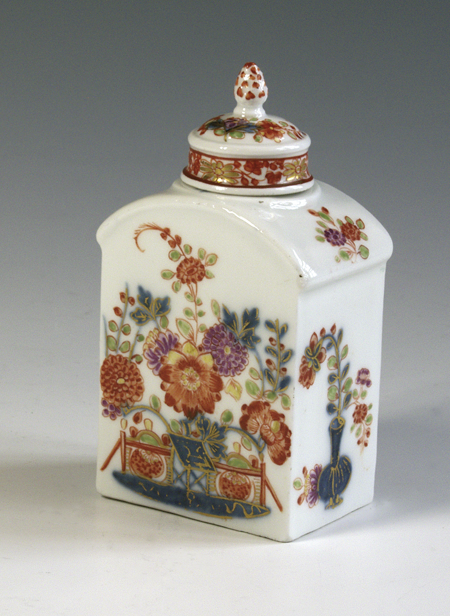 Herbatnica, Saksonia, Miśnia, 1735 – 1740; porcelana, farby naszkliwne i kobalt podszkliwnie, złocenia; wys. 12,7 cm, szer. 6,7 cm, gł. 4,8 cm; zbiory Muzeum Zamoyskich w Kozłówce, nr inw. MPK/CS/20 Fotografia przedstawia porcelanowy pojemnik na herbatę w kształcie prostopadłościanu, o łukowato zakreślonych krawędziach górnych, z przykrywką. W kolorze białym z dekoracją w typie imari. Przykrywka zwieńczona szyszką, w części środkowej lekko wyniesiona, na wybrzuszeniu bukiet drobnych kwiatów, niżej obwiedziona taśmą z ornamentem kwiatowym. Na szerszej ściance motyw stylizowanego płotu i stolika, przy nich kwiaty chryzantem i peonii. Na węższej ściance waza z kwiatami.