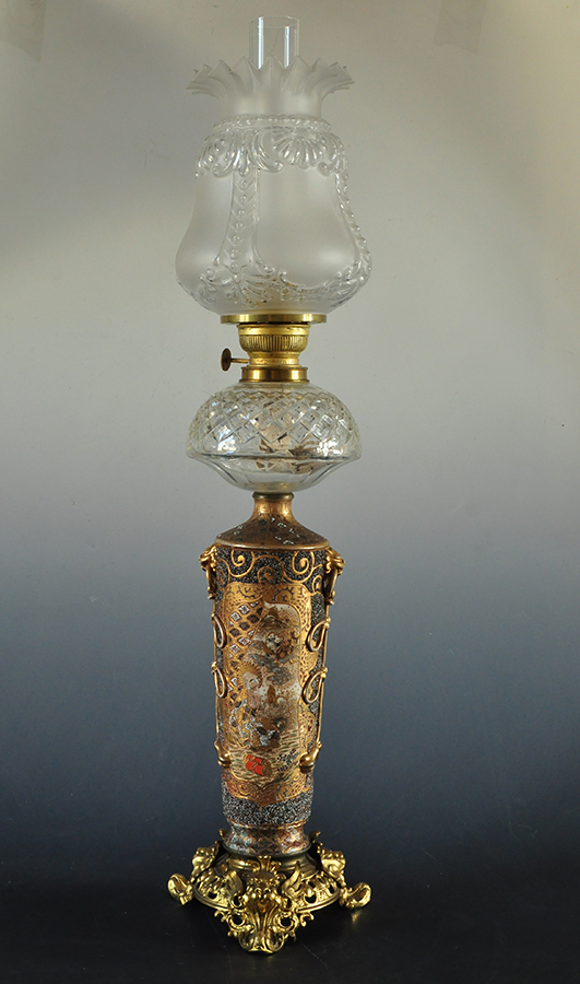 Lampa naftowa stołowa, Japonia, Niemcy, Satsuma, Berlin, przełom XIX/XX wieku; nr inw. MPK/CS/2051 Lampa o mosiężnej podstawie, na trzech nogach zwieńczonych maskami uskrzydlonych lwów. Kamionkowy korpus jest walcowaty i smukły, z bardzo bogatą dekoracją w kolorystyce z przewagą złota, bieli i różu. Po bokach korpusu ziarnista faktura. Klosz z matowego szkła zwieńczony kryzą. Po bokach plastyczne uchwyty z głowami lwów. Pojemnik na naftę szklany, półkulisty, szlifowany w ukośną kratę. Palnik pierścieniowy.