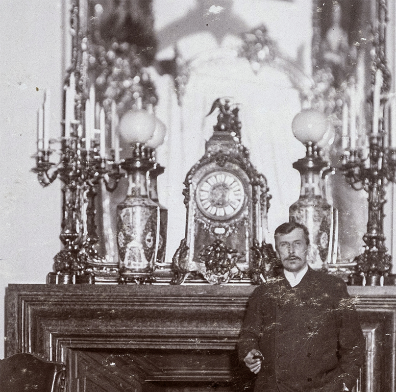 Fragment fotografii ukazującej Konstantego Zamoyskiego w Salonie Czerwonym pałacu w Kozłówce lata 90. XIX wieku, fotografia zabytkowa, nr inw. MPK/F/210 Zabytkowa czarno-biała fotografia ukazująca fragment wnętrza pomieszczenia. Przy kominku stoi postawny, elegancko ubrany mężczyzna-Konstanty Zamoyski. W wysuniętej dłoni trzyma cygaro. Włosy ułożone na prawą stronę, uformowane wąsy. Za nim, sięgający wysokością jego ramion marmurowy kominek, na którym ustawiony jest centralnie udekorowany złoceniami i figurą Chronosa zegar. Po obu stronach zegara ustawione są lampy naftowe o porcelanowych podstawach i kulistych kloszach, obok nich wysokie kandelabry. Za dekoracją, nad kominkiem wysokie lustro.