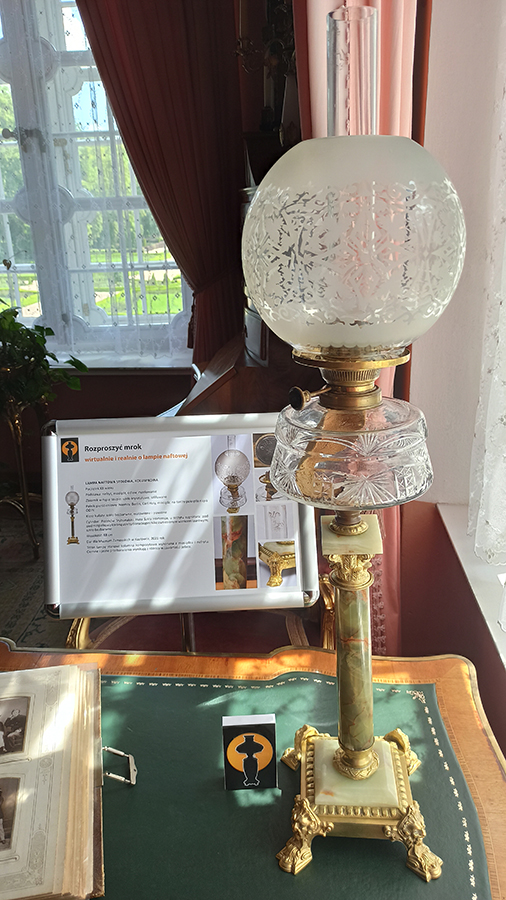 Lampa naftowa kolumnowa na biurku w Gabinecie południowym pałacu kozłowieckiego Na biurku lampa o oszronionym szklanym kloszu i zielonej, wykonanej z nefrytu podstawie, w postaci kolumienki. Zbiornik na naftę wykonany jest ze rżniętego szkła. W tle widoczna tablica z informacją na temat lampy, obok lampy niewielki znaczek przedstawiający na czarnym tle nimb światła i czarną sylwetę lampy naftowej. W tle okno z białą firana i zasłoną w odcieniu różu.