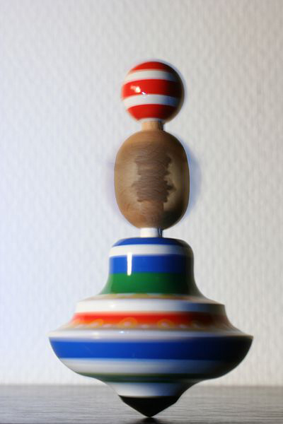 Bączek, zabawka, źródło: https://pl.wiktionary.org/wiki/toupie#/media/Plik:Spinning_top_-_prague.jpg Fotografia ukazuje ceramiczną zabawkę – bąka wprawianą w ruch obrotowy dookoła swojej osi. Dolna część ma kształt spłaszczonej cebuli zakończonej szpicem, malowanej w pasy w kolorze niebieskim, zielonym i czerwonym, kolejny element podłużny w kolorze drewna, zwieńczenie stanowi kulka w czerwono białe pasy.