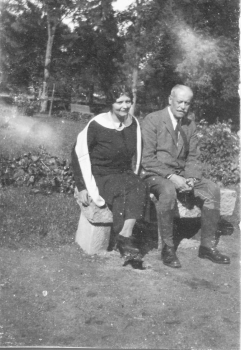 5. Maria i Adam Zamoyscy, Kozłówka, lata 20. XX w. Fotografia archiwalna, czarno-biała, wykonana w latach dwudziestych XX wieku, przedstawia Marię Zamoyską i jej męża, Adama Zamoyskiego. Kobieta i mężczyzna siedzą na kamiennej ławce. Kobieta ma ciemne włosy, spięte z tyłu, ubrana jest w ciemną suknię z białym kołnierzem i biały szal. Mężczyzna ma siwe krótkie włosy i wąsy, ubrany jest w szary garnitur i jasną koszulę z krawatem.