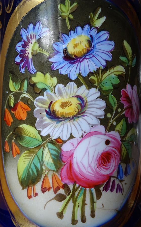 Fotografia kolorowa przedstawia fragment porcelanowego korpusu lampy naftowej. W owalu bukiet kwiatów w kolorach białym, różowym, niebieskim, pomarańczowym. Pomiędzy kwiatami zielone liście.