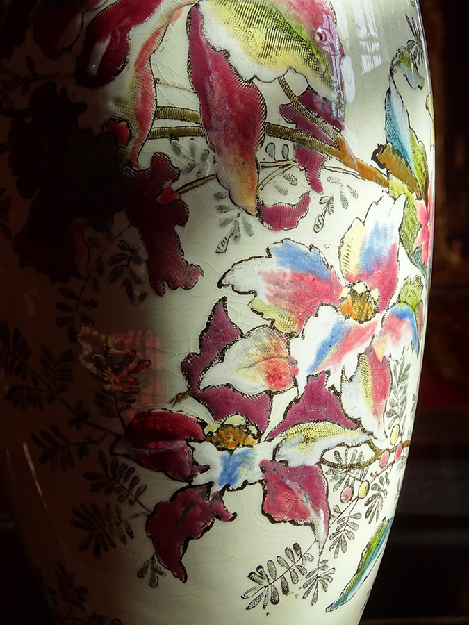 Fotografia kolorowa przedstawia lampę naftową o metalowej podstawie, na której osadzony jest owoidalny porcelanowy korpus ozdobiony kwiatami katlei i liściastymi gałązkami. Kolorystyka dekoracji jest w odcieniach różu, błękitu i żółci.