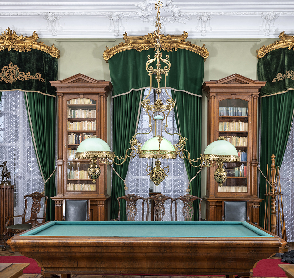 Kolorowa współczesna fotografia ukazująca wnętrze Biblioteki w pałacu kozłowieckim. Na pierwszym planie potrójna, mosiężna lampa naftowa o trzech szklanych kloszach z zielonego mlecznego szkła, zawieszona nad stołem bilardowym. W tle widoczne trzy okna, nad którymi są złocone, dekorowane ornamentami karnisze i do nich zamocowane aksamitne, ciemno – zielone lambrekiny