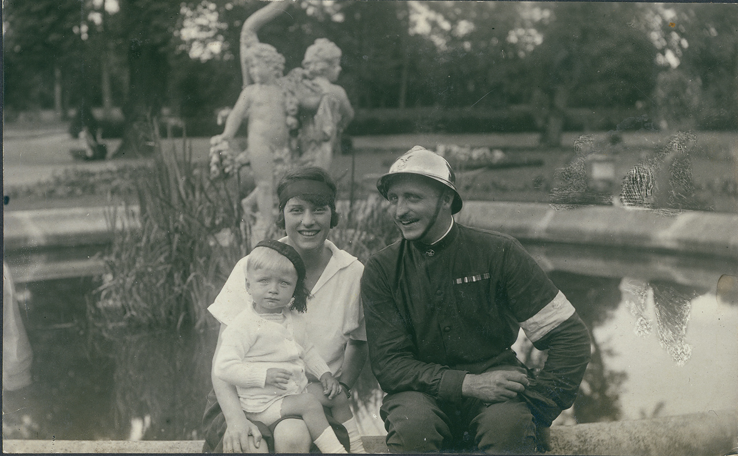 1.	Fotografia archiwalna z lat 30. XX wieku przedstawia Jadwigę i Aleksandra Zamoyskich z synem Adamem, siedzących na brzegu fontanny w parku kozłowieckim. Uśmiechnięta młoda kobieta w białej sukience i z czarną opaską we włosach. Trzyma na kolanach kilkuletniego chłopca w białym ubranku, białych skarpetkach i ciemnym nakryciu głowy. Po jej prawej stronie uśmiechnięty mężczyzna w ciemnym stroju i błyszczącym hełmie. W tle fontanna z rzeźbą – dwa amorki stojące tyłem do siebie. W głębi park. Fotografia czarno-biała.