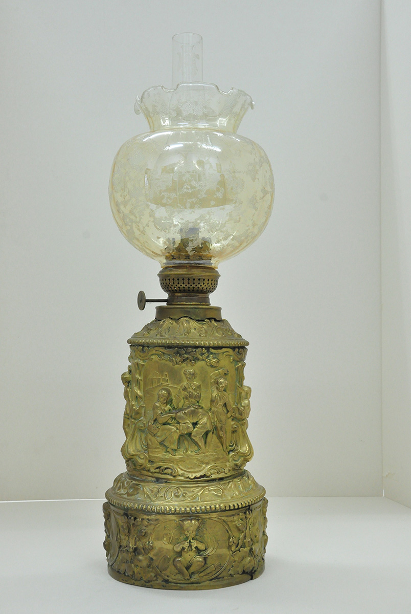 Lampa naftowa, stołowa dekorowana potrójnie powtórzoną sceną klapsa, Francja (?), XIX wiek; mosiądz, wytłaczanie, kucie, odlew, szkło lodowe, barwione, cylinder: Polska, Piotrków Trybunalski, Huta Szkła „Hortensja”; szkło bezbarwne; nr. inw. MPK/ZM/1415 Fotografia kolorowa przedstawiająca XIX-wieczną mosiężną lampę naftową, o cylindrycznym korpusie, z półplastyczną dekoracją figuralną i ornamentalną. Na wysokiej okrągłej podstawie, w regularnie powtórzonych czterech medalionach w roślinnej oprawie, reliefowe frontalnie ukazane putta grające na instrumentach. Wokół nieco węższego korpusu trzykrotnie powielona scena rodzajowa (wymierzania kary cielesnej lub zabawy w salonowca). W kwaterach ujętych bordiurą z kwiatów i ornamentów na tle oddalonej wyniesionej architektury, siedząca postać kobieca. Na jej kolanach spoczywa głowa chłopca lub mężczyzny pochylonego przy niej. Dwaj inni stojący z tyłu, jeden z uniesioną ręką, ubrani w surduty z wywiązanymi koszulami. Pomiędzy tymi głównymi częściami oraz przy palniku spadziste pasy dekoracji wypełnione wicią roślinną, z perełkowaniem przy brzegu. Klosz lampy jest kulisty, pękaty, tulipanowy, z falistą kryzą.