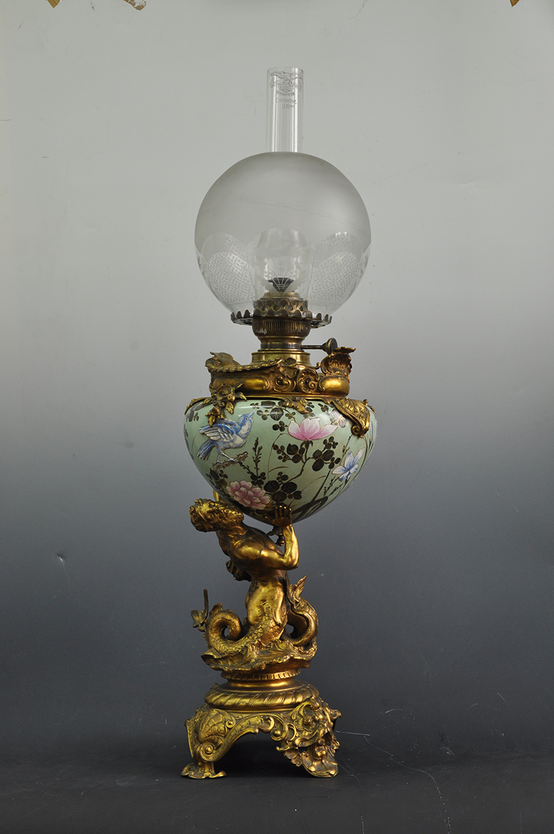 Lampa naftowa stołowa, salonowa z majolikowym zbiornikiem podtrzymywanym przez pełnoplastyczną figurę Trytona, Niemcy (?), Austria (?), koniec XIX wieku; mosiądz, majolika, odlew, cyzelowanie, farby naszkliwne, nr inw. MPK/ZM/1402 Fotografia kolorowa przedstawiająca lampę naftową z końca XIX wieku, z majolikowym malowanym korpusem podtrzymywanym przez mosiężnego Trytona oraz z kulistym szklanym kloszem. Podstawa utworzona z połączonych ornamentów, z czterema nóżkami. Postać boga morskiego Trytona (górna część ciała człowieka, dolna w kształcie ryby) przedstawiona jest z głową odchyloną do tyłu i skręconą w bok; pokryty łuskami rybi ogon zakończony jest rozdwojoną płetwą. Rękoma uniesionymi do góry podtrzymuje zbiornik lampy. Czasza półowoidalna, jasnozielonej barwy, z malowaną częściowo wypukłą dekoracją: gałązki japońskiej wiśni (sakura), gałązki zbliżone wyglądem do krzewów różanych, ulistnione łodygi z kwiatami przypominającymi lilie, delikatnie zarysowane cienkie trawki. Po obu stronach na gałązkach półplastyczne biało-niebieskie ptaszki, wraz z elementami roślinnymi obwiedzione złotymi konturami. Czasza ujęta obręczą utworzoną z ornamentów. Klosz kulisty, w górnej części szkło zmatowione, w dolnej przezroczyste z wytrawioną dekoracją w polach zamkniętych łukami.