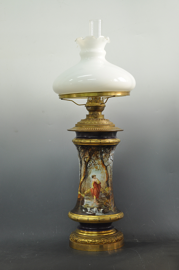 Lampa naftowa stołowa, salonowa z dekoracją malarską, Anglia, wytwórnia George’a Jonesa, dekoracja malarska Brickman, ok. 1872 roku, mosiądz, odlew, cyzelowanie, złocenie, porcelana, farby naszkliwne, szkło bezbarwne, szkło mleczne, nr. inw. MPK/CS/2052 Fotografia kolorowa przedstawiająca XIX-wieczną lampę naftową stołową, salonową z dekoracją malarską przedstawiającą leśny pejzaż z postacią młodej kobiety w strumyku. Podstawa lampy wykonana jest z mosiądzu i ozdobiona wieńcem laurowym. Korpus w kształcie zbliżonym do cylindrycznego, na węższej, kobaltowej i złoconej stopie na dole i u góry, ze złoconym półwałkiem dekorowanym drobnym ornamentem. Na korpusie namalowany leśny pejzaż ze strumykiem i niskim wodospadem oraz młodą kobietą w pomarańczowo-żółtej szacie, wchodzącą do wody. Zbiornik mosiężny, zdobiony perełkowaniem, pierścieniem z liści laurowych i rozetowo ułożonymi wokół palnika płatkami. Klosz ze szkła mlecznego, kopulasty, z falistą kryzą.