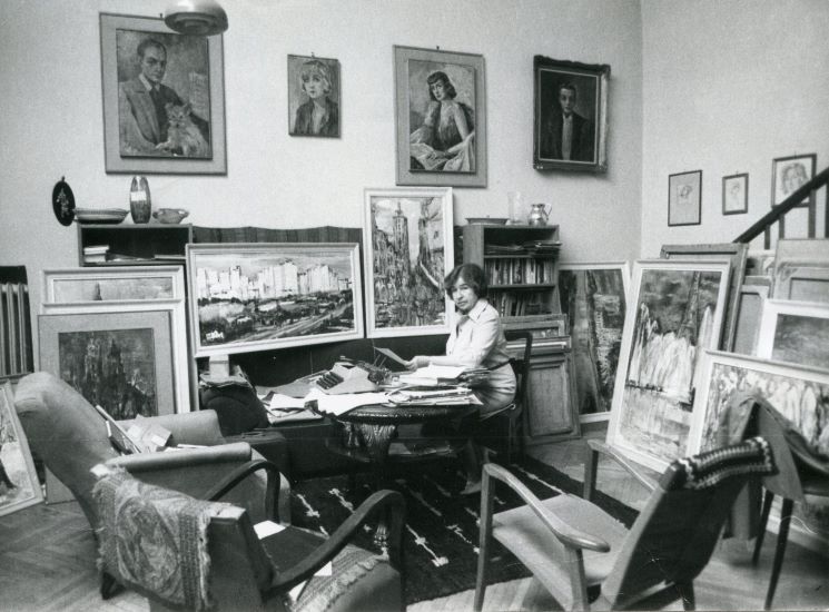 Zdjęcie artystki w jej pracowni. Wilimowska siedzi w fotelu przy dużym okrągłym stole, przy którym stoją jeszcze trzy puste fotele. Na stole piętrzą się papiery, dokumenty, stoi maszyna do pisania. Na ścianach oraz przy ścianach obrazy w ramach. Całość w stylistyce lat 60.