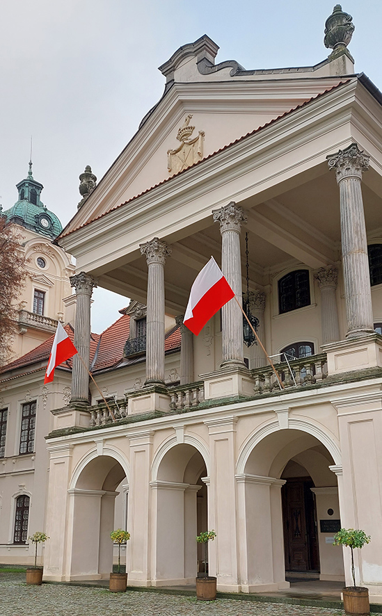 Fotografia kolorowa przedstawiająca fasadę pałacu. Na tarasie dwie biało-czerwone flagi.