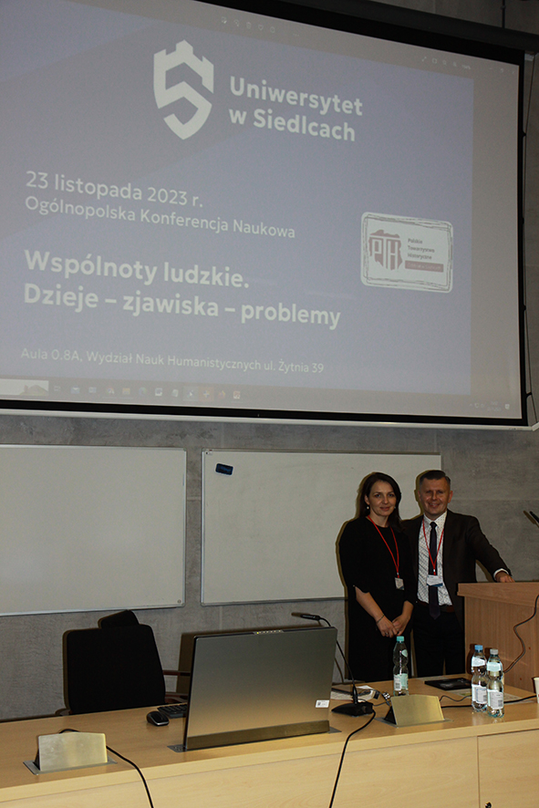 zdjęcie współczesne, przedstawiające kobietę i mężczyznę na tle ekranu multimedialnego na którym logo i nazwa Uniwersytetu w Siedlcach oraz logo Polskiego Towarzystwa Historycznego oraz napis: „23 listopada 2023 r. Ogólnopolska Konferencja Naukowa Wspólnoty ludzkie. Dzieje – zjawiska – problemy”. Na pierwszym planie fragment stołu konferencyjnego.