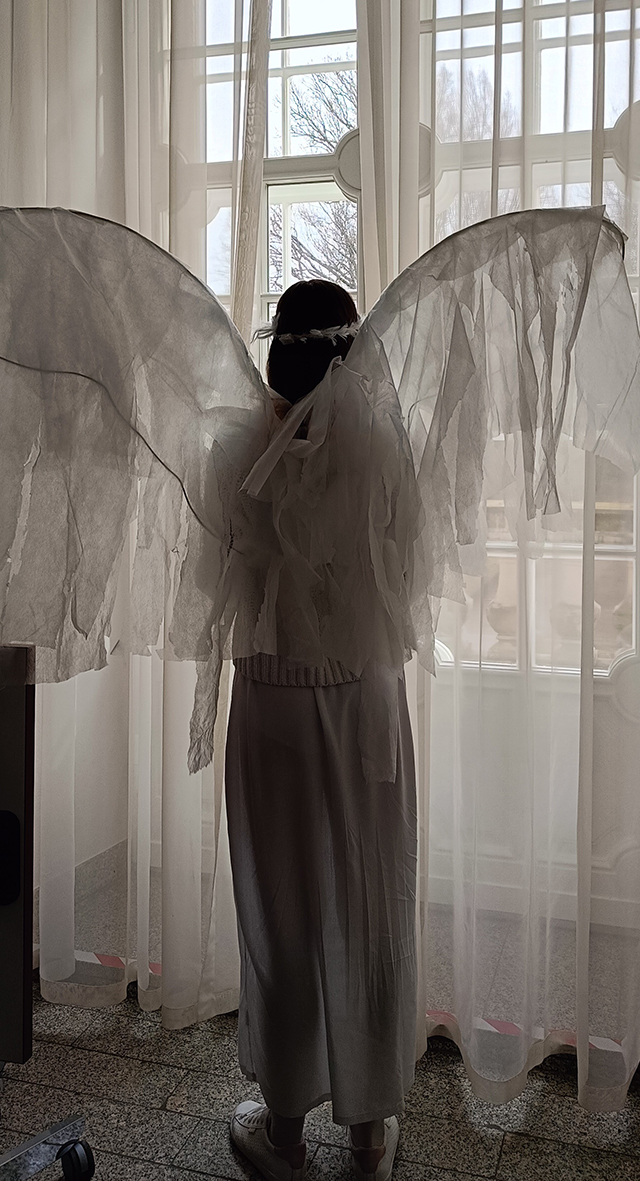 Kobieta ubrana w zwiewny strój anioła wygląda przez duże okno, w którym zawieszone są firany z delikatnego, prześwitującego materiału.