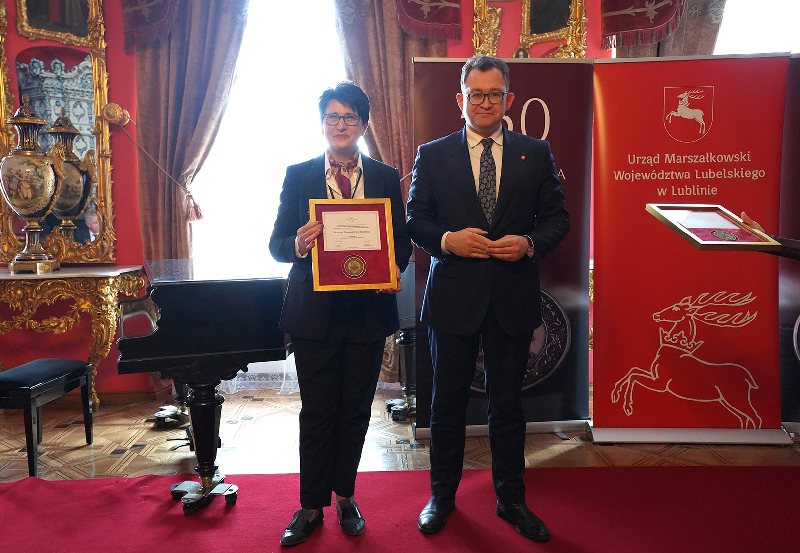 4. Uroczyste Otwarcie Sezonu Kulturalnego 2024 w Lubelskich Muzeach Samorządowych Na zdjęciu widzimy panią dyrektor Muzeum Zamoyskich w Kozłówce, która dumnie trzyma w dłoniach przyznany medal. Jej uśmiech na twarzy świadczy o głębokim zadowoleniu. Obok niej stoi Członek Zarządu Województwa Lubelskiego, pan Bartłomiej Bałaban. W tle widoczne są charakterystyczne elementy wnętrza pałacowego, dopełniające uroczystego klimatu tego wyjątkowego wydarzenia.