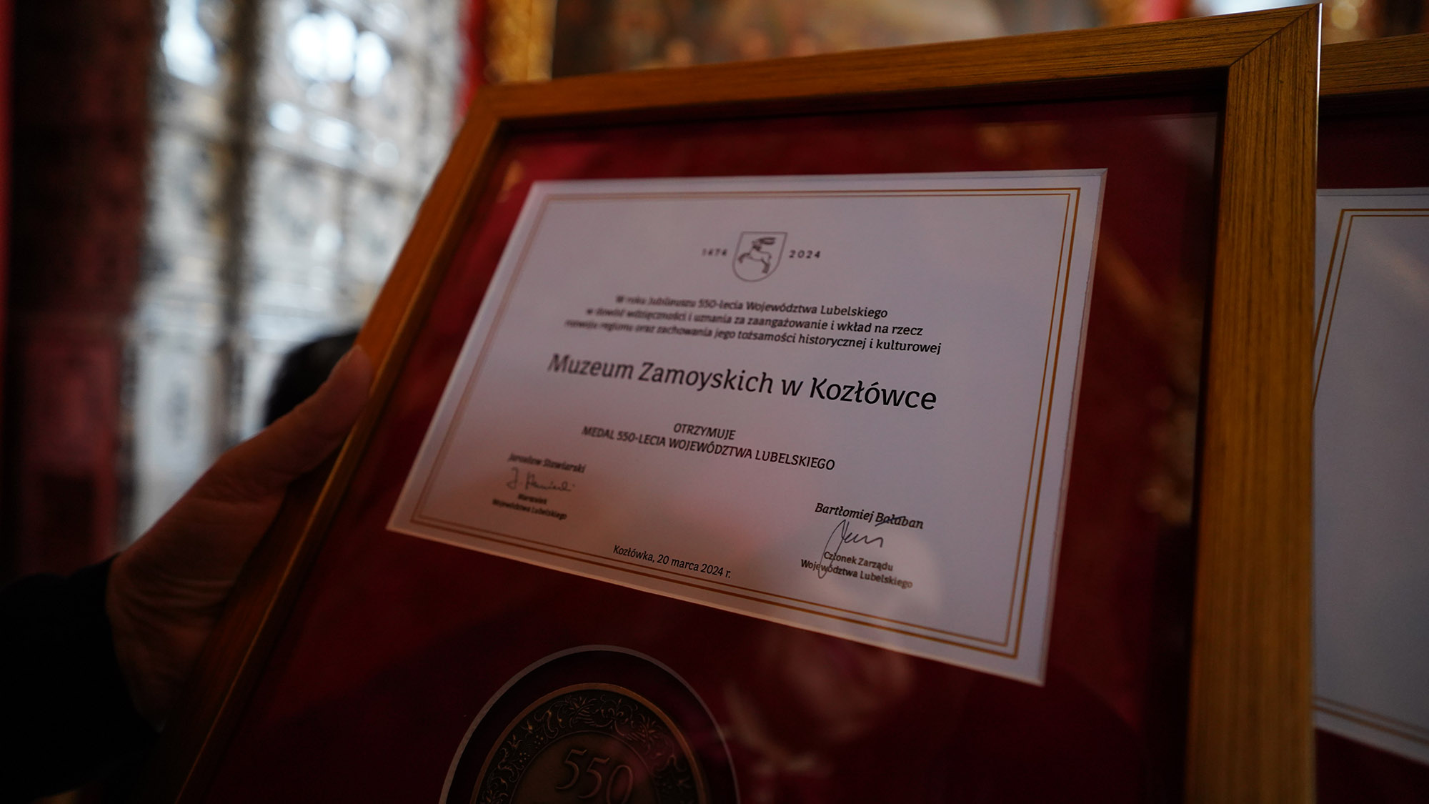 6. 4. Uroczyste Otwarcie Sezonu Kulturalnego 2024 w Lubelskich Muzeach Samorządowych Na zdjęciu ozdobny medal przyznany Muzeum Zamoyskich w Kozłówce w wyrazie uznania i wdzięczności za zaangażowanie wkład na rzecz rozwoju regionu i zachowania jego tożsamości historycznej i kulturowej. Medal w raz z uzasadnieniem został oprawiony w elegancką ramkę. Jego powierzchnia wydaje się lśnić lekko w świetle, co dodaje mu elegancji.