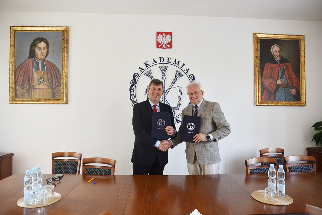 Na zdjęciu widzimy dwóch mężczyzn stojących na środku eleganckiego pomieszczenia, najprawdopodobniej gabinetu rektorskiego Akademii Zamojskiej. Są to Andrzej Pruszkowski, p.o. Dyrektora Muzeum Zamoyskich w Kozłówce oraz Paweł Skrzydlewski, profesor Akademii Zamojskiej. Obaj uśmiechają się i ściskają sobie dłonie, trzymając jednocześnie ciemnoniebieskie teczki z logo oraz napisem „REKTOR”, co sugeruje podpisanie oficjalnej umowy lub porozumienia.
Mężczyzna po lewej ma na sobie czarny garnitur, jasnoniebieską koszulę i czerwony krawat. Mężczyzna po prawej ubrany jest w jasnoszarą kraciastą marynarkę, białą koszulę i krawat w odcieniach granatu. Obaj mają siwe włosy, przy czym mężczyzna po lewej krótsze i ciemniejsze.
W tle widnieje herb Polski z orłem w koronie, pod nim okrągłe logo z nazwą „AKADEMIA” i elementami przypominającymi berła rektorskie. Po obu stronach logo wiszą dwa portrety historycznych postaci w złotych ramach – z lewej strony postać w czerwono-brązowym stroju z krzyżem maltańskim, z prawej mężczyzna z białą brodą w czerwonym żupanie, trzymający szablę.
Przed mężczyznami stoi długi drewniany stół z krzesłami, na którym znajdują się butelki wody, okulary, długopis i szklanki.