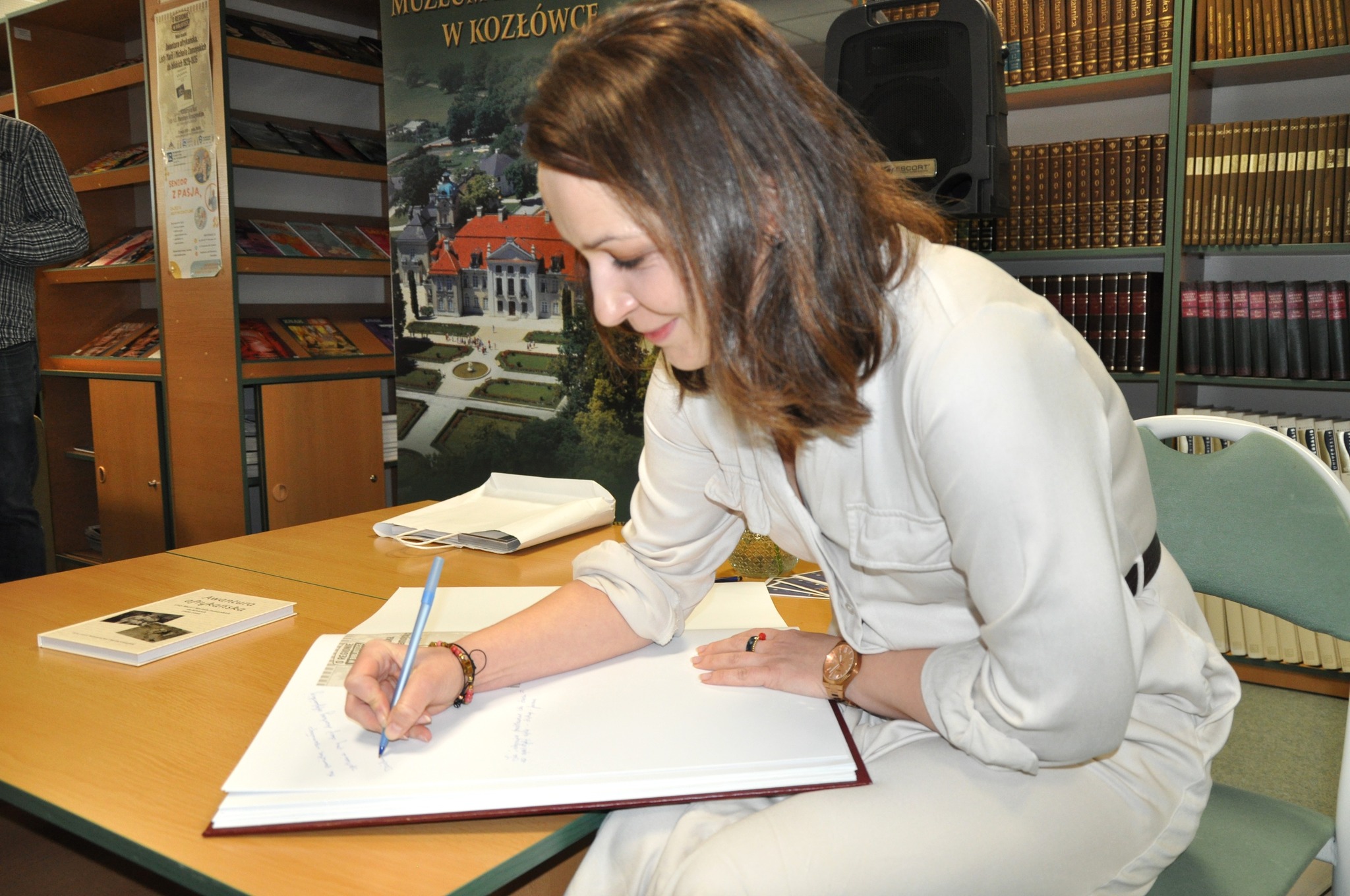 Spotkanie wokół książki “Afrykańska awantura. Listy Marii i Michała do bliskich 1929-1935” w Książnicy Zamojskiej w Zamościu Na zdjęciu widzimy tę samą kobietę, która wcześniej prowadziła spotkanie. Siedzi przy stole, pochylona nad dużym, otwartym albumem – prawdopodobnie księgą pamiątkową. Pisze coś długopisem, uśmiechając się lekko. Ubrana jest w jasny kombinezon z podwiniętymi rękawami, na ręce ma zegarek i kilka bransoletek. W tle – regały z książkami oraz plakat promujący Muzeum Zamoyskich w Kozłówce, przedstawiający pałac i ogród.