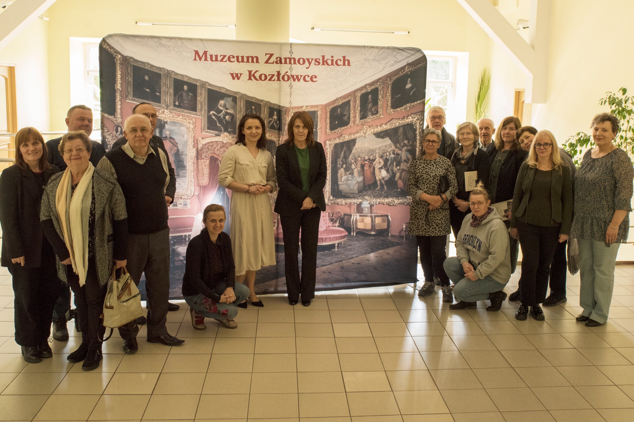 Spotkanie wokół książki “Afrykańska awantura. Listy Marii i Michała do bliskich 1929-1935” w Książnicy Zamojskiej w Zamościu Grupa około dwudziestu osób stoi przed dużym banerem przedstawiającym wnętrze pałacu z czerwonymi ścianami i bogatymi zdobieniami – to zapewne wnętrza Muzeum Zamoyskich w Kozłówce. W centrum zdjęcia stoją dwie kobiety – jedna w jasnej sukience (ta sama autorka co na poprzednich zdjęciach), obok niej druga kobieta w czarnym garniturze i zielonej bluzce. Pozostali uczestnicy otaczają je z obu stron. Większość osób jest ubrana w stonowane kolory, patrzą w stronę aparatu, część uśmiechnięta.