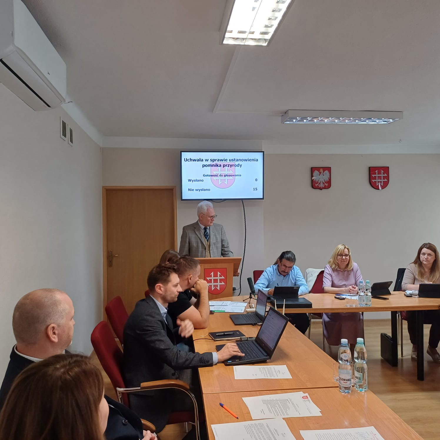 Dąb szypułkowy z przypałacowego parku Pomnikiem Przyrody Zdjęcie przedstawia salę obrad Rady Miejskiej, prawdopodobnie w Kamionce, podczas oficjalnego posiedzenia. Na pierwszym planie widać duży, drewniany stół konferencyjny w kształcie litery „L”, przy którym siedzi kilkoro radnych – zarówno kobiety, jak i mężczyźni – korzystających z laptopów i dokumentów.
Na dalszym planie, za mównicą z herbem gminy, stoi starszy mężczyzna z siwymi włosami, w jasnej marynarce i krawacie – najprawdopodobniej przewodniczący lub prowadzący obrady. Nad jego głową, na ścianie, znajduje się ekran z wyświetlonym tytułem głosowanego punktu:
„Uchwała w sprawie ustanowienia pomnika przyrody”.
Poniżej widać liczby:
– „Głos oddano: 0”,
– „Wyjęto kart do głosowania: 0”,
– „Nie wyjęto: 15”.
Po bokach ekranu wiszą dwa godła – biały orzeł w czerwonym tle (godło Polski) oraz herb gminy Kamionka.
W sali panuje atmosfera skupienia, a wygląd wnętrza – białe ściany, drewniana podłoga, prosty wystrój – wskazuje na typowe miejsce pracy samorządowej.