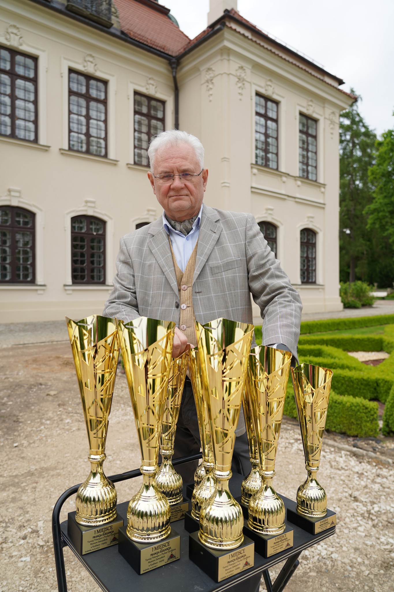 Dni kultury francuskiej. Francja Elegancja Na zdjęciu widzimy starszego mężczyznę stojącego na zewnątrz przed eleganckim, klasycystycznym budynkiem o jasnej elewacji i ciemnych oknach z ozdobnymi szprosami. Budynek ma wysoki dach pokryty dachówką i bogato zdobione fasady. Otoczenie to zadbany ogród z przyciętymi krzewami i żwirową alejką.
Mężczyzna ma siwe włosy i okulary, ubrany jest w elegancki, jasnoszary garnitur w kratę, pod którym nosi beżowy sweter i błękitną koszulę. Ma także apaszkę lub szalik pod szyją, co dodaje mu wytwornego wyglądu.
Przed nim na czarnym wózku ustawionych jest dziewięć dużych, złotych pucharów. Puchary mają smukły kształt z ozdobnymi wycięciami i czarne podstawy z tabliczkami. Na tabliczkach widać napis „1 MIEJSCE” i inne informacje, świadczące o tym, że są to trofea przyznawane za zajęcie pierwszego miejsca w jakimś konkursie lub zawodach. Mężczyzna trzyma jeden z pucharów i patrzy poważnie w stronę aparatu.
Całość kompozycji sugeruje oficjalną okazję, być może uroczystość wręczenia nagród lub wydarzenie związane z prestiżowym konkursem.