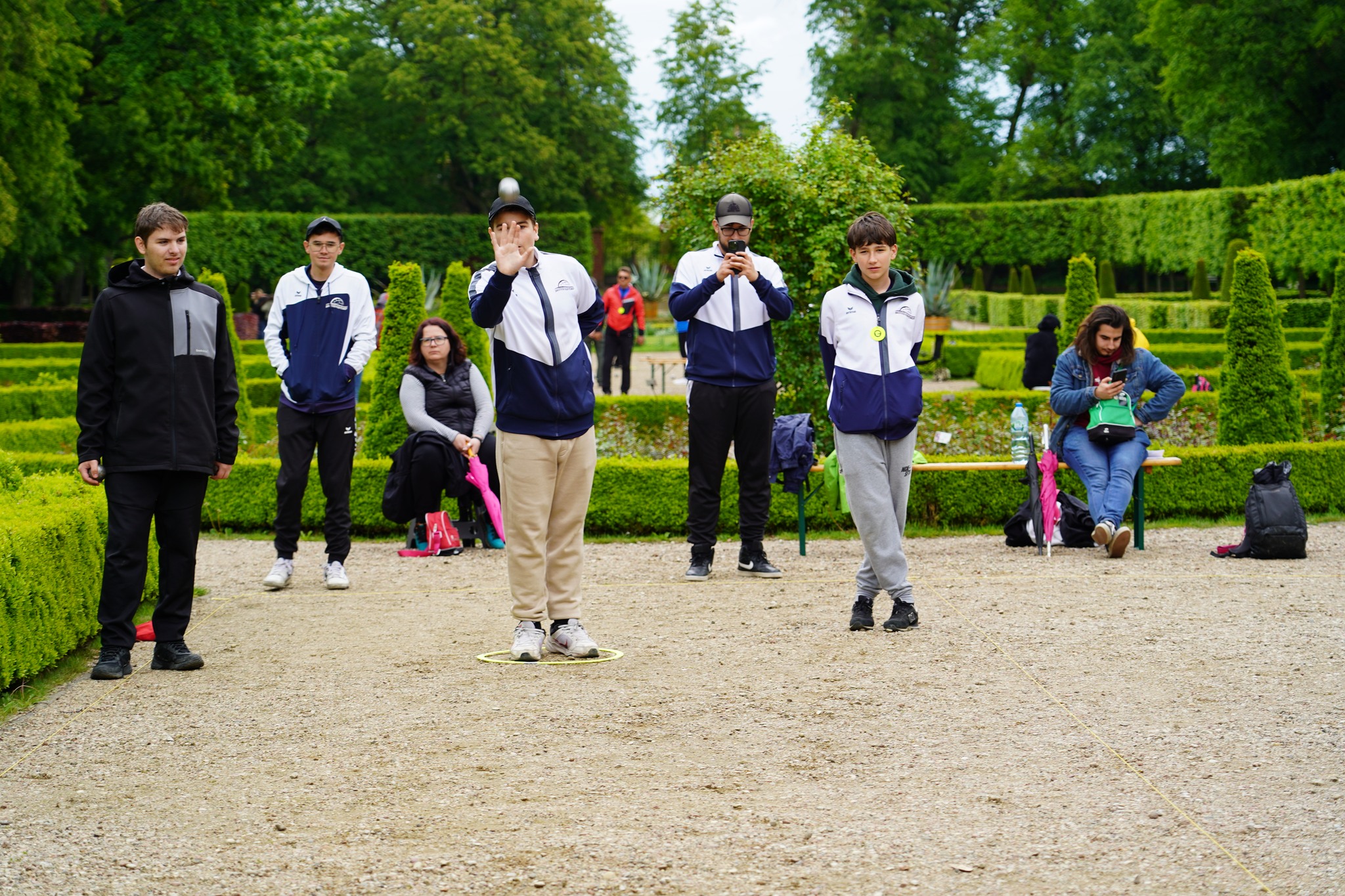 Dni kultury francuskiej. Francja Elegancja Na zdjęciu przedstawiona jest scena z rozgrywek sportowych na świeżym powietrzu, odbywających się w eleganckim ogrodzie w stylu francuskim, otoczonym precyzyjnie przyciętymi żywopłotami i alejkami żwirowymi. Tło stanowią zielone krzewy i wysokie drzewa.
W centrum kadru znajduje się młody chłopak w jasnych spodniach i biało-granatowej kurtce sportowej z daszkiem na głowie. Znajduje się w wyznaczonym żółtym kółku na ziemi i właśnie rzuca kulą – wygląda na to, że uczestniczy w grze w pétanque (bule). Jego dłoń jest uniesiona w geście rzutu, a wzrok skupiony przed siebie.
Wokół niego znajduje się kilku innych młodych ludzi, również w sportowych strojach, prawdopodobnie członków tej samej drużyny. Jeden z nich robi zdjęcie lub film telefonem, inny przygląda się grze z boku. W tle, na ławkach, siedzą kibice i uczestnicy – część z nich odpoczywa, rozmawia lub korzysta z telefonów.
Atmosfera jest spokojna, skoncentrowana, a otoczenie sugeruje, że wydarzenie odbywa się w prestiżowym lub historycznym miejscu.