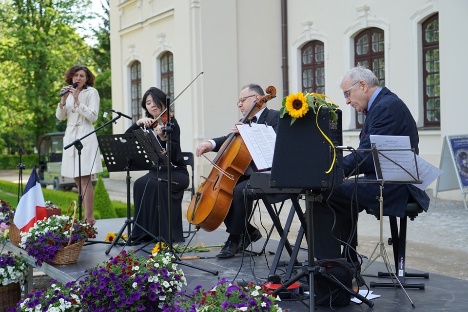 Dni kultury francuskiej. Francja Elegancja Na zdjęciu widzimy występ muzyczny odbywający się na zewnętrznej scenie, ustawionej na tle eleganckiego, jasnego budynku w stylu pałacowym. Scena udekorowana jest kolorowymi kwiatami w wiklinowych koszach oraz małą flagą Francji, co może sugerować francuski charakter wydarzenia lub międzynarodowy kontekst.
Na scenie występuje kwartet:
Po lewej stronie stoi wokalistka w kremowym płaszczu/trenczu, trzymająca mikrofon i śpiewająca z zaangażowaniem.
Obok niej siedzi skrzypaczka ubrana w elegancką, czarną suknię, skupiona na grze i odczytująca nuty z pulpitu.
Dalej widzimy wiolonczelistę, również elegancko ubranego, grającego na instrumencie z nut przed sobą.
Po prawej stronie siedzi pianista przy elektronicznym pianinie ozdobionym słonecznikami, pochylony nad klawiaturą i skoncentrowany na grze.
Za artystami znajduje się elegancka fasada budynku z dużymi, stylowymi oknami i dekoracyjnymi obramowaniami. Otoczenie sugeruje, że koncert odbywa się w ogrodzie pałacowym lub na dziedzińcu willi, w otoczeniu zieleni i kwiatów. Atmosfera jest uroczysta, kameralna i artystyczna.