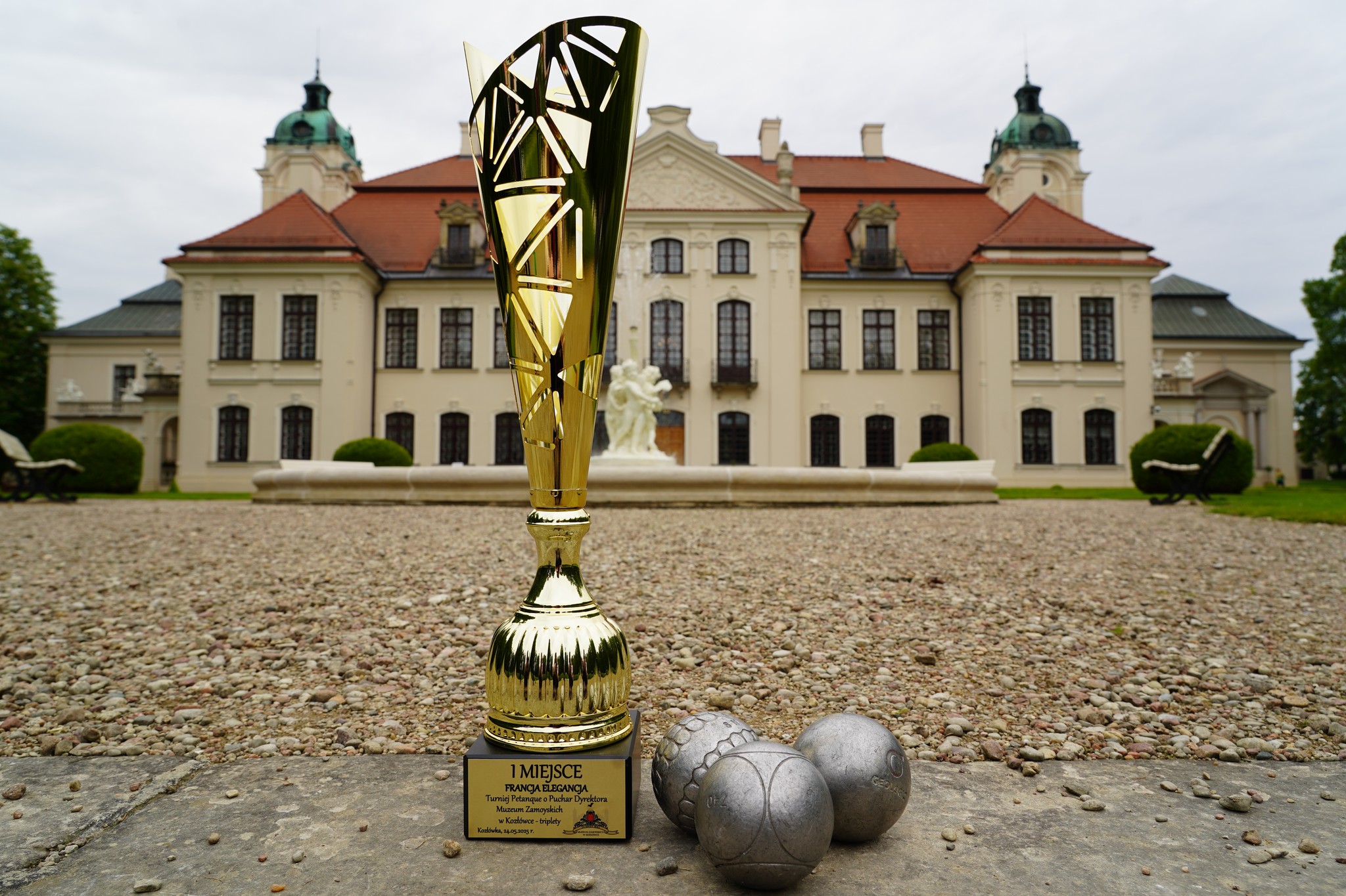 Dni kultury francuskiej. Francja Elegancja Na zdjęciu widzimy elegancki, złoty puchar ustawiony na pierwszym planie, stojący na żwirowej alei przed dużym, klasycystycznym pałacem z czerwonym dachem i dwiema wieżami. U stóp pucharu znajdują się trzy metalowe kule do gry w petankę (boules), co sugeruje, że zdjęcie dokumentuje zakończenie lub sukces w turnieju tej dyscypliny.
Na podstawie pucharu widnieje napis:
„1 MIEJSCE – FRANCJA ELEGANCJA
Turniej Pétanque o Puchar Dyrektora Muzeum Zamoyskich
w Kozłówce – drużyny”
Kozłówka, 25.05.2024 r.”
W tle znajduje się wspomniany pałac, będący siedzibą Muzeum Zamoyskich w Kozłówce. Centralnie widać także fontannę z białą rzeźbą, dodającą majestatycznego charakteru tej scenerii. Pogoda jest pochmurna, ale jasna, co pozwala dostrzec wyraźnie wszystkie detale architektoniczne i elementy kompozycji. Zdjęcie emanuje elegancją, tradycją i prestiżem wydarzenia.