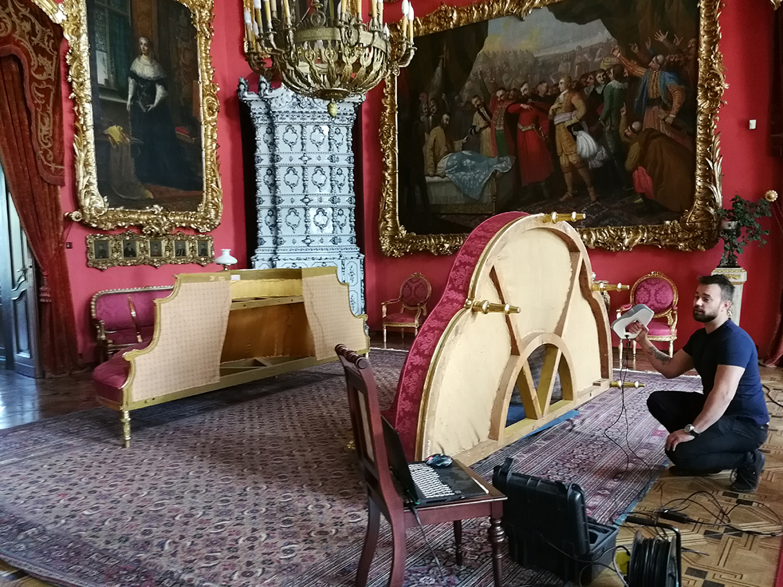Skanowanie kanapy, tzw. konfidentki Na kolorowej fotografii wnętrze dużego salonu. Pośrodku na podłodze duży dywan we wzory geometryczne w kolorze bordowym, białym i niebieskim. Na pierwszym planie, nieco po prawej krzesło, na nim laptopem, obok otwarta skrzynka na sprzęt i kable. Za krzesłem półokrągła duża kanapa, z siedziskiem w kształcie trójliścia, położona siedziskiem do góry, prawym bokiem do widza. Tapicerka w kolorze bordowym, z motywem kwiatowym, podbicie i oskrzynienie w kolorze jasnym, cztery widoczne nóżki złocone. Na prawo od kanapy mężczyzna z aparaturą do skanowania w prawej dłoni. Na lewo od kanapy druga kanapa o takim samym kształcie stojąca tyłem do widza. W głębi, od lewej, fragment otwartych drzwi przesłoniętych lambrekinem i kotarą w kolorze bordowo-złotym. Obok, , duży portret stojącej kobiety w stroju XVI-wiecznym, obraz w bogato rzeźbionych złoconych ramach. Pod portretem 5 małych portretów w podobnych ramach. Dalej, na prawo, w rogu salonu, wysoki piec ze zdobnymi kaflami w kolorze biało-niebieskim. Obok, na ścianie duży obraz ze sceną zbiorową (mężczyzna siedzący na łóżku w otoczeniu żywo gestykulujących mężczyzn). Obraz w bogato rzeźbionych złoconych ramach. Pod nim dwa krzesła z tapicerką w bordowym kolorze i złotym oskrzynieniem. Pośrodku obrazu, od góry fragment mosiężnego żyrandola.