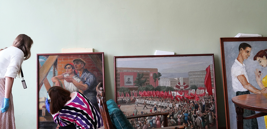 Muzeum Zamoyskich w Kozłówce partnerem wystawy „Lato, które zmieniło wszystko. Festiwal 1955” w Muzeum Warszawy Na zdjęciu widzimy trzy obrazy ustawione pionowo pod ścianą w pomieszczeniu o jasnozielonej ścianie. Obrazy są oprawione w proste, drewniane ramy. Przed nimi znajdują się dwie kobiety – jedna stoi po lewej stronie, pochylając się w stronę obrazu, druga klęczy przodem do obrazu i fotografuje go telefonem. Obie kobiety wykonują czynności ostrożnie – jedna z nich ma na dłoniach niebieskie rękawiczki ochronne.
Opis obrazów:
Pierwszy obraz z lewej przedstawia młodą kobietę i mężczyznę w robotniczym stroju, stojących razem przy rusztowaniu z cegieł. Kobieta opiera się o mężczyznę, oboje patrzą z nadzieją w dal. Styl realistyczny, typowy dla sztuki socrealistycznej.
Środkowy obraz ukazuje wielką manifestację z czerwonymi sztandarami. Tłum ludzi maszeruje przez miasto, niosąc transparent z napisem „OBROŃMY POKÓJ”. W tle znajduje się ogromny gmach z wielkim portretem Stalina oraz napisem w języku polskim, który głosi: „ZJEDNOCZONA PARTIA ROBOTNICZA”. To typowa scena propagandowa z lat 50., ukazująca entuzjazm tłumów wobec władzy.
Obraz po prawej stronie pokazuje młodą parę – chłopaka i dziewczynę – w czułym, intymnym geście. Dziewczyna w żółtej sukience pochyla się w kierunku chłopaka, który wręcza jej kwiat. Scena utrzymana w pastelowych barwach, bardziej subtelna i osobista niż pozostałe dwa dzieła.
Na pierwszym planie, z prawej strony, widać fragment drewnianego stołu oraz oparcie zielonego, stylowego fotela lub kanapy.
Całość sceny sprawia wrażenie przygotowań do ekspozycji – prawdopodobnie obrazy są właśnie katalogowane lub dokumentowane przed wystawą.