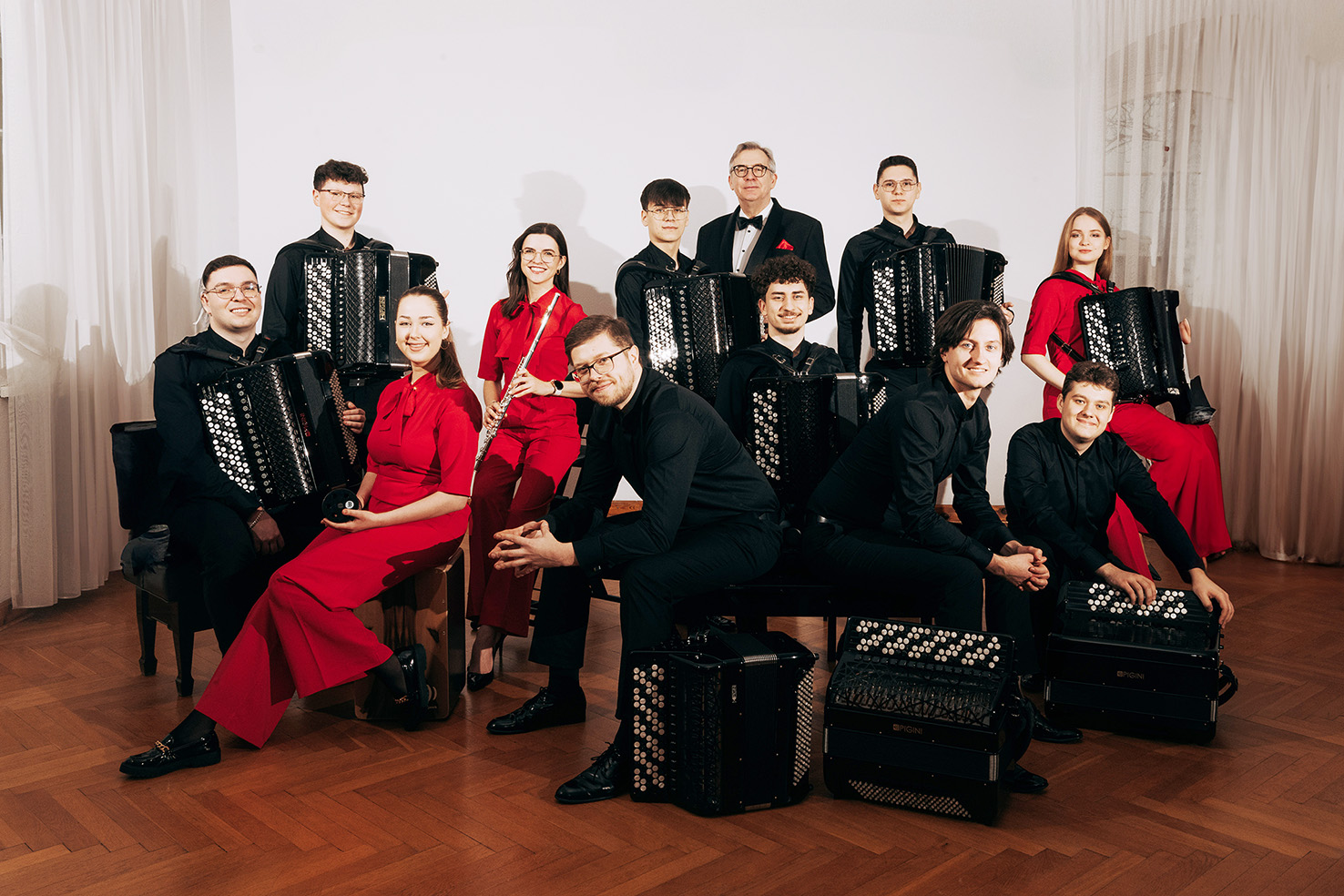 Koncert Kameralnej Orkiestry Akordeonowej Arti Sentemo Na fotografii widzimy Kameralną Orkiestrę Akordeonową Arti Sentemo – grupę jedenastu młodych muzyków oraz ich dyrygenta. Zdjęcie wykonano we wnętrzu – pomieszczenie ma jasne ściany i drewnianą podłogę w jodełkę. Z lewej i prawej strony widoczne są białe, prześwitujące zasłony.
Muzycy siedzą i stoją w dwóch rzędach, patrząc prosto w obiektyw i uśmiechając się delikatnie. Ubrani są elegancko, w jednolitym stylu: mężczyźni głównie w czarnych koszulach lub garniturach, kobiety w czerwonych, klasycznych strojach. Jeden z członków zespołu (mężczyzna w środku, z siwymi włosami i okularami) wyróżnia się – ma na sobie smoking z muchą i czerwony ozdobny element w kieszonce marynarki, co sugeruje, że może być dyrygentem lub opiekunem zespołu.
Większość muzyków trzyma w rękach duże, czarne akordeony koncertowe z białymi guzikami, ustawione pionowo na kolanach. Jedna z kobiet (druga od lewej, w czerwonym stroju) trzyma flet poprzeczny, co wskazuje na obecność instrumentów towarzyszących w składzie orkiestry. Na twarzach wszystkich wykonawców widać skupienie i pozytywną energię.
Zespół został ustawiony starannie i symetrycznie, co tworzy wrażenie profesjonalizmu i zgrania. Zdjęcie emanuje ciepłem, harmonią i pasją do muzyki.