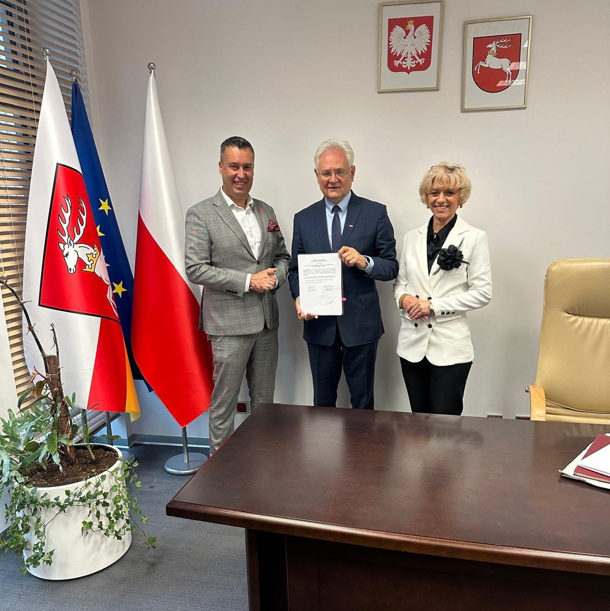 Rozstrzygnięcie konkursu na Dyrektora Muzeum Zamoyskich w Kozłówce Na zdjęciu widzimy trzy osoby stojące w eleganckim biurze na tle flag i godła.
W centrum kadru stoi mężczyzna w ciemnym garniturze, trzymający w rękach dokument skierowany przodem do kamery – to prawdopodobnie Uchwała Zarządu Województwa. Z jego wyrazu twarzy można wyczytać powagę i satysfakcję.
Po jego lewej stronie (z prawej strony patrzącego) stoi kobieta w białym garniturze z czarnym kwiatem przypiętym do klapy marynarki. Uśmiecha się serdecznie w stronę obiektywu.
Po prawej stronie mężczyzny (z lewej strony patrzącego) stoi inny mężczyzna – w szarym garniturze, z ciemnymi włosami i również uśmiechnięty. Lekko dotyka ramienia osoby w centrum i gestykuluje dłonią, jakby właśnie złożył gratulacje.
W tle widoczne są:
– trzy flagi (flaga województwa lubelskiego, flaga Polski i flaga Unii Europejskiej),
– dwa godła na ścianie: orzeł biały (godło Polski) i herb województwa lubelskiego – biały jeleń na czerwonym tle.
Po lewej stronie kadru widoczna jest roślina doniczkowa, a po prawej duże biurko z dokumentami.
Całość utrzymana jest w oficjalnym, reprezentacyjnym tonie – to wyraźnie moment wręczenia ważnego dokumentu w instytucji publicznej.
