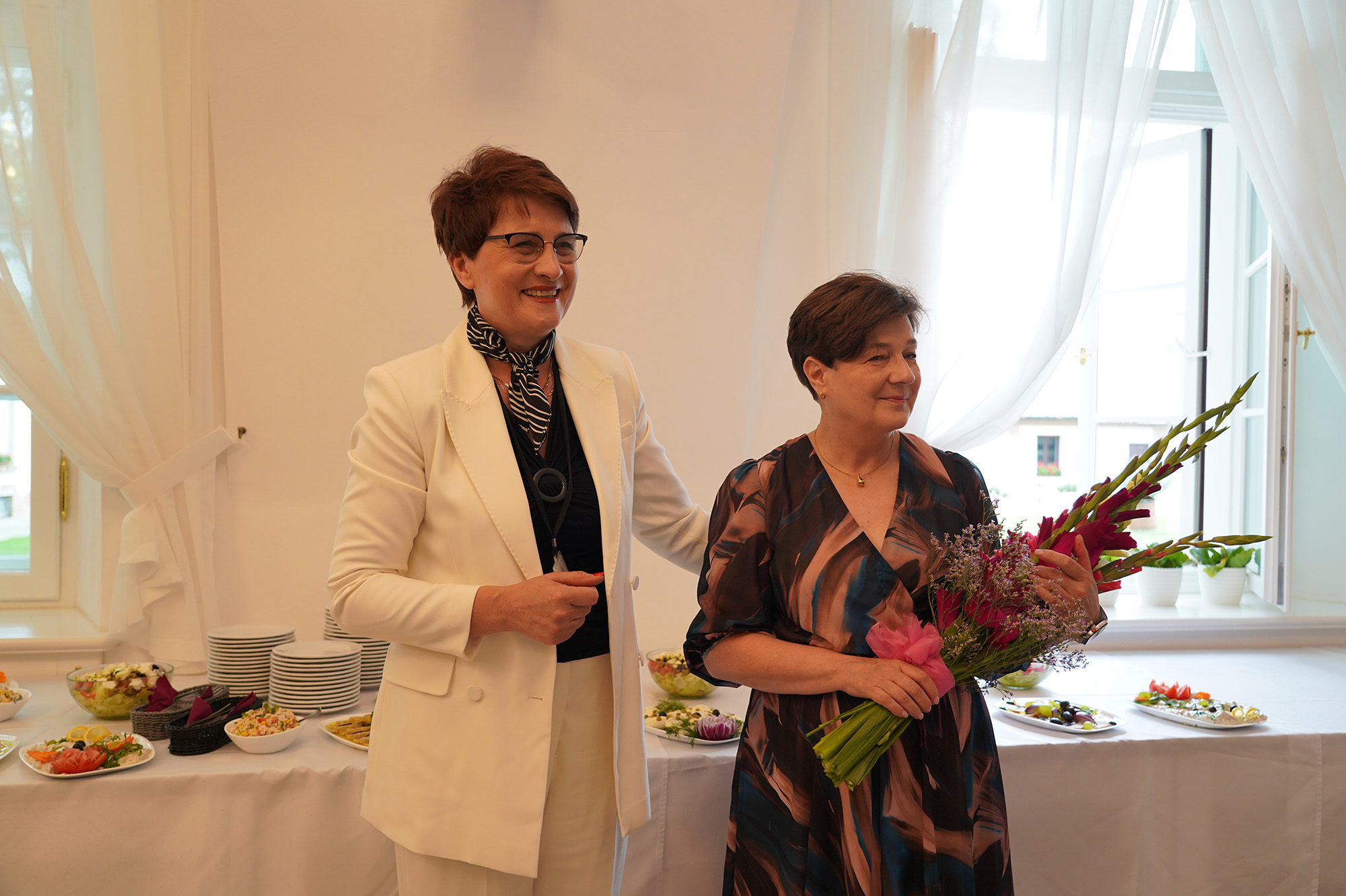 fot_2.jpg Zdjęcie przedstawia uroczystość z okazji przejścia na emeryturę p. Elżbiety Dybały. Centralną postacią jest p. Elżbieta, kobieta w średnim wieku stojąca na tle stołu, trzymająca w rękach bukiet różowych i fioletowych mieczyków. Obok niej, stoi p. Dyrektor Bożena Ćwiek, ubrana w biały garnitur. Obie kobiety są uśmiechnięte.