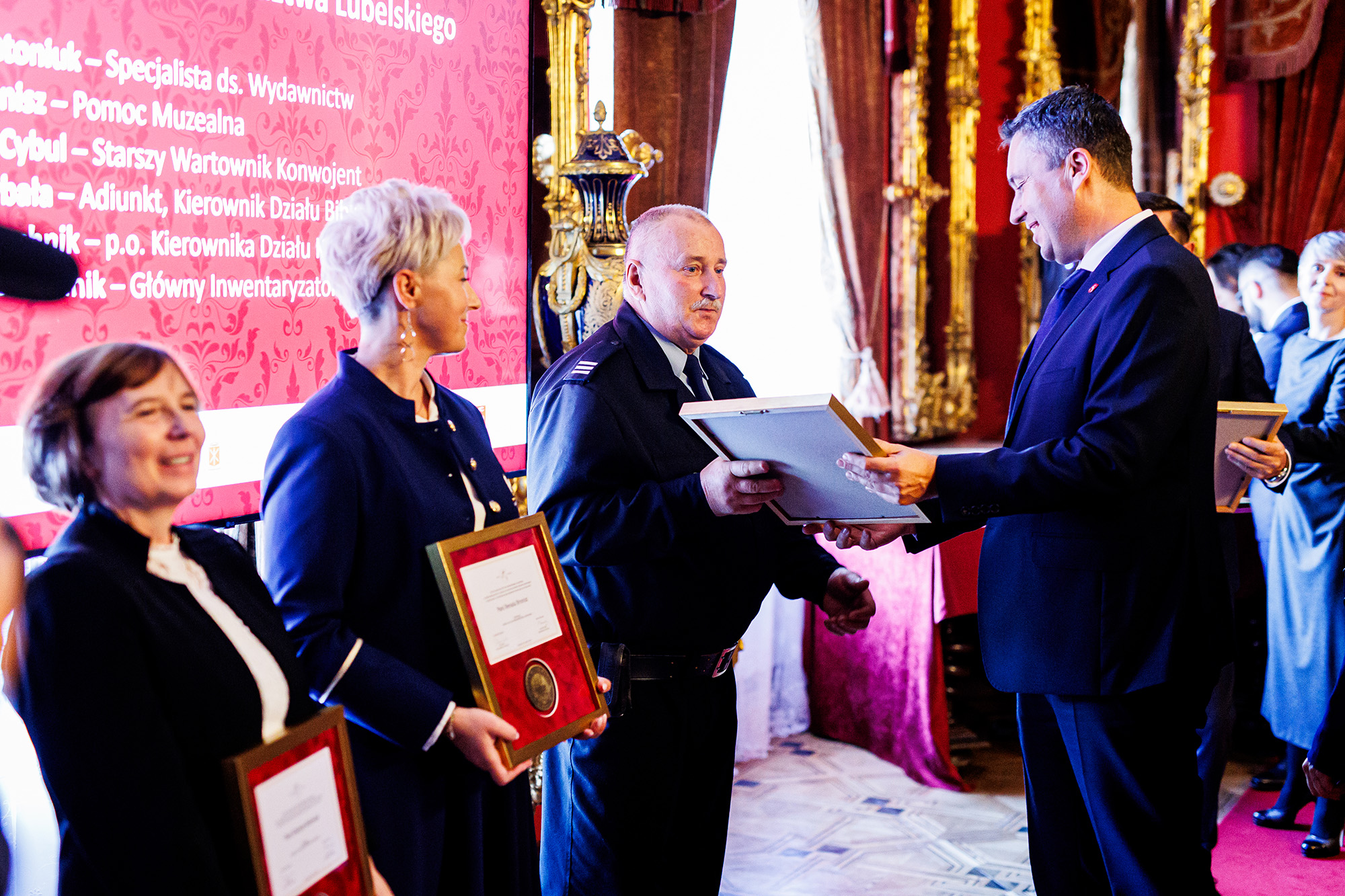 fot_3.jpg Opis ogólny:
Fotografia przedstawia uroczystość wręczania odznaczeń w eleganckim, zabytkowym wnętrzu – sali reprezentacyjnej pałacu w Kozłówce. Na pierwszym planie widocznych jest kilka osób, które otrzymują dyplomy i wyróżnienia. Atmosfera jest oficjalna i uroczysta.
Postaci:
Po prawej stronie kadru znajduje się mężczyzna w garniturze, p. Marcin Szewczak, członek Zarządu Województwa Lubelskiego, który wręcza nagrodę starszemu mężczyźnie w mundurze, p. Waldemarowi Cyblowi. Ten drugi mężczyzna przyjmuje dyplom z lekkim uśmiechem, trzymając go oburącz.
Obok nich, z lewej strony, stoją dwie kobiety ubrane elegancko w ciemne żakiety. Obie trzymają już odebrane dyplomy w ramkach – każda z nich prezentuje je z dumą. Jedna z kobiet ma krótko ścięte, blond włosy.
W tle po prawej stronie widać więcej osób w kolejce po odbiór wyróżnień.
Tło i otoczenie:
Uroczystość odbywa się w bogato zdobionym wnętrzu z czerwonymi zasłonami, złotymi zdobieniami oraz ciemnymi meblami. W głębi po lewej stronie widoczna jest duża plansza z tekstem na czerwonym tle, prawdopodobnie przedstawiająca nazwiska wyróżnionych osób lub nazwę wydarzenia.
Atmosfera:
Oficjalna, uroczysta i pełna szacunku. Widać, że jest to ważna chwila dla uczestników, być może związana z wieloletnią pracą lub zasługami.