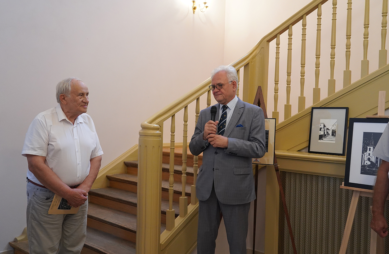 Uroczyste otwarcie wystawy, od lewej r Ireneusz Targoński oraz Dyrektor Muzeum Andrzej Pruszkowski Zdjęcie przedstawia uroczyste otwarcie wystawy w oficynie północnej Muzeum Zamoyskich w Kozłówce. Na zdjęciu dwaj mężczyźni stoją w eleganckim wnętrzu przy schodach. Po lewej stronie znajduje się mężczyzna ubrany w jasną koszulę z krótkim rękawem i jasne spodnie. Trzyma broszurę lub katalog, spogląda w stronę drugiego mężczyzny. To autor wystawy Ireneusz Targoński. Po prawej stronie stoi drugi mężczyzna w szarym garniturze, niebieskiej koszuli i krawacie z delikatnym wzorem. Ma białe włosy i okulary. Trzyma mikrofon i przemawia. To Dyrektor Muzeum Andrzej Pruszkowski. Za nim znajduje się żółta klatka schodowa z drewnianymi poręczami. Obok niego ustawione są sztalugi z oprawionymi grafikami – jedna z nich przedstawia architekturę z ostrymi kontrastami czerni i bieli, inna pokazuje budynek z wieżą kościelną. Tło stanowi jasna ściana i drewniane schody prowadzące w górę.