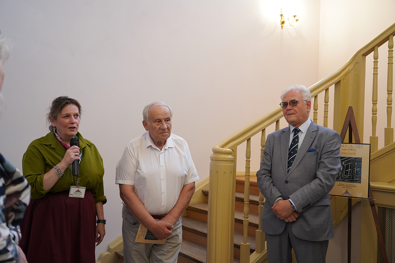 Uroczyste otwarcie wystawy, od lewej koordynatorka wystawy Magdalena Pilsak, Ireneusz Targoński oraz Dyrektor Muzeum Andrzej Pruszkowski Zdjęcie przedstawia uroczyste otwarcie wystawy w oficynie północnej Muzeum Zamoyskich w Kozłówce. W centrum kadru znajduje się troje ludzi, stojących na tle jasnej klatki schodowej z drewnianą poręczą w kolorze jasnego brązu lub beżu. Pomieszczenie o kremowych ścianach jest dobrze oświetlone światłem naturalnym oraz kinkietami. Po lewej stronie stoi kobieta o jasnej cerze i brązowych włosach spiętych z tyłu. Ubrana jest w zieloną bluzkę z podwiniętymi rękawami oraz bordową, rozkloszowaną spódnicę. Na szyi ma różową apaszkę, a na szyi zawieszony identyfikator. Mówi do trzymanego w ręku mikrofonu. To koordynatorka wystawy Magdalena Pilsak. W środku stoi mężczyzna o siwych włosach. Ubrany jest w białą, koszulę z krótkimi rękawami oraz beżowe spodnie. W rękach trzyma złożony folder lub broszurę. To autor wystawy Ireneusz Targoński. Po prawej stronie stoi kolejny mężczyzna, z białymi włosami, w okularach. Ma na sobie elegancki, jasnoszary garnitur z krawatem w ciemne pasy i białą koszulę. Na piersi ma niebieską poszetkę. To Dyrektor Muzeum Andrzej Pruszkowski. W tle, obok schodów, stoi sztaluga z plakatem wystawy. Widoczna jest część tekstu i grafiki, ale szczegóły są niewyraźne.